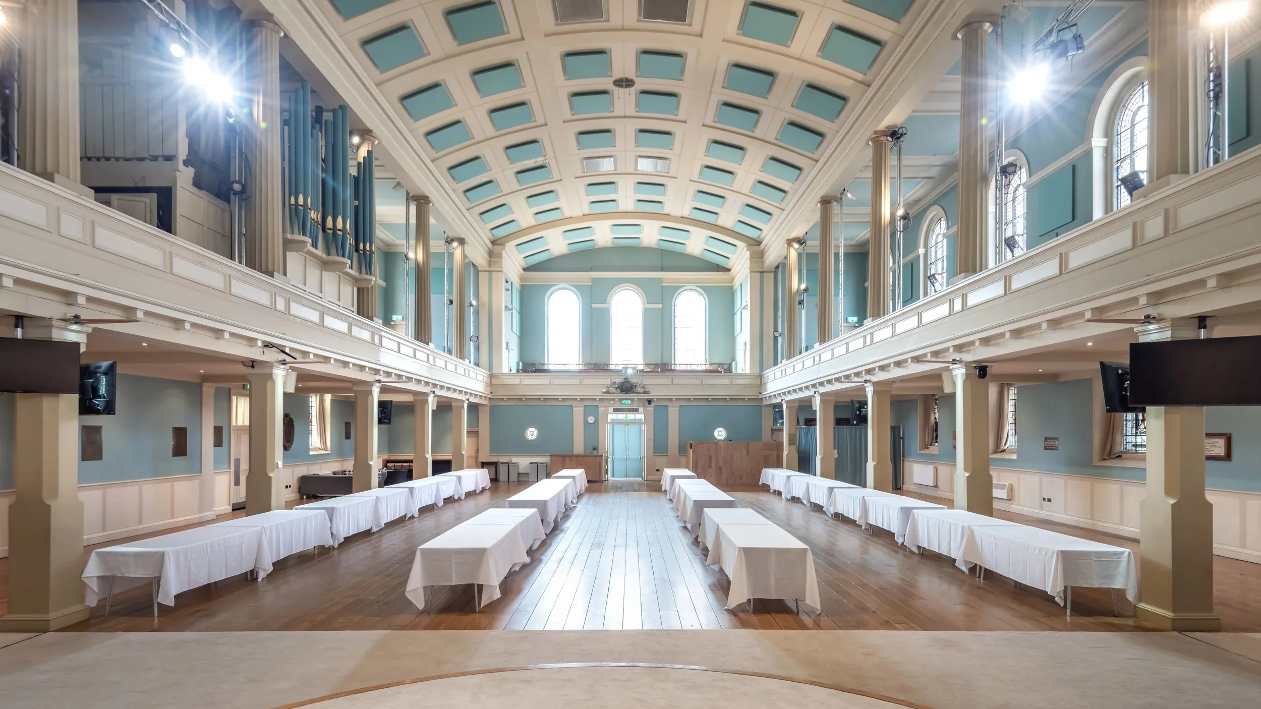 An elegant, spacious hall with high arched windows, light blue walls, a decorative vaulted ceiling, and rows of white-covered tables set up for an event or banquet.