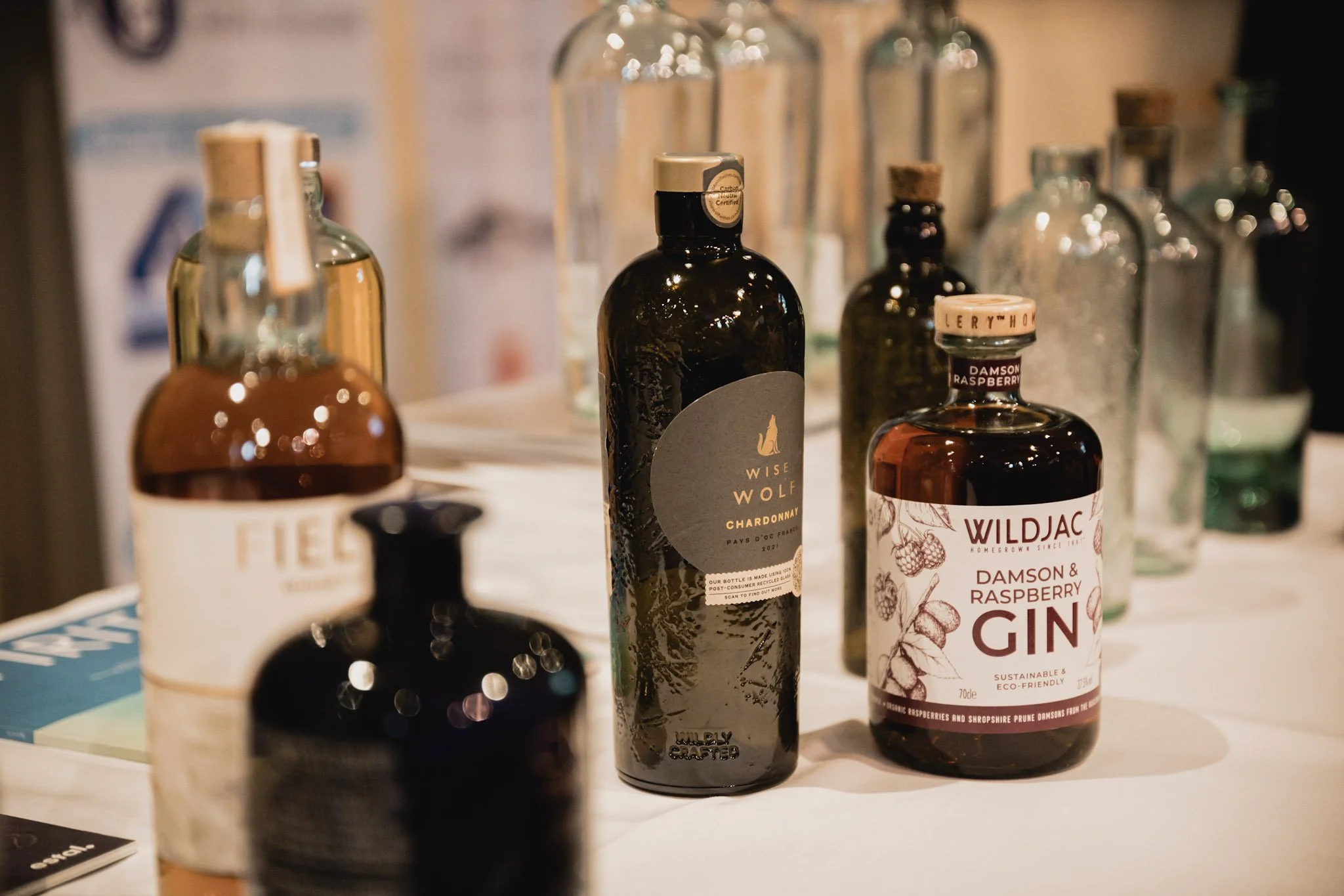 A collection of bottles including Wise Wolf Chardonnay, WildJAC Damson & Raspberry Gin, and other assorted bottles on a table.