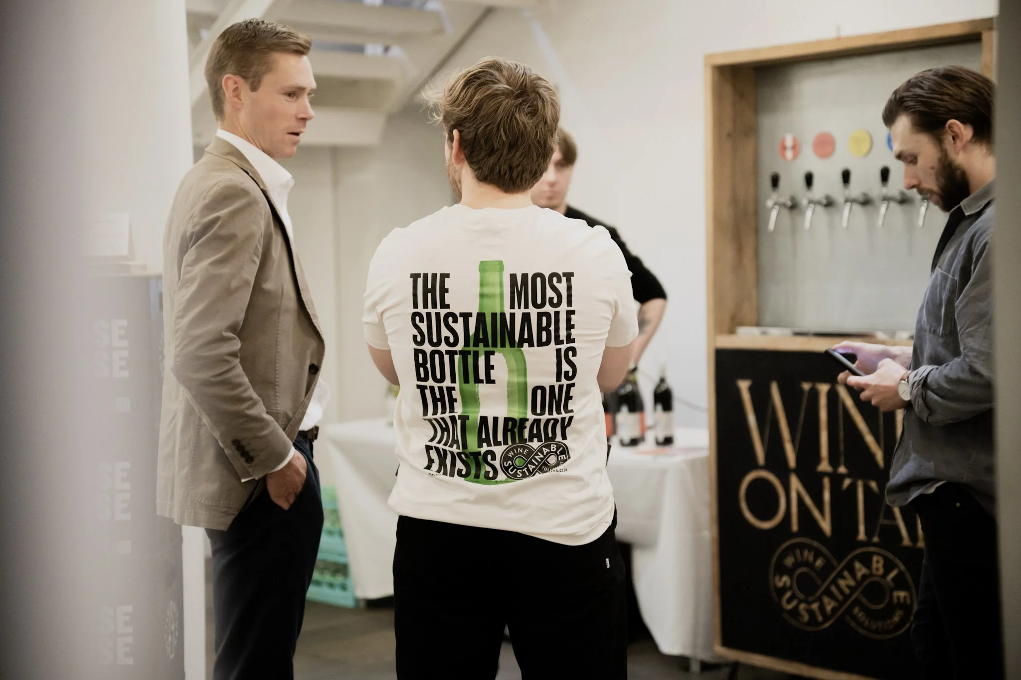 Three men are standing and conversing at a booth promoting sustainable wine bottles. One man is wearing a beige blazer, another has a white T-shirt with a message about sustainable bottles, and the third man is in a dark gray shirt using his phone. B