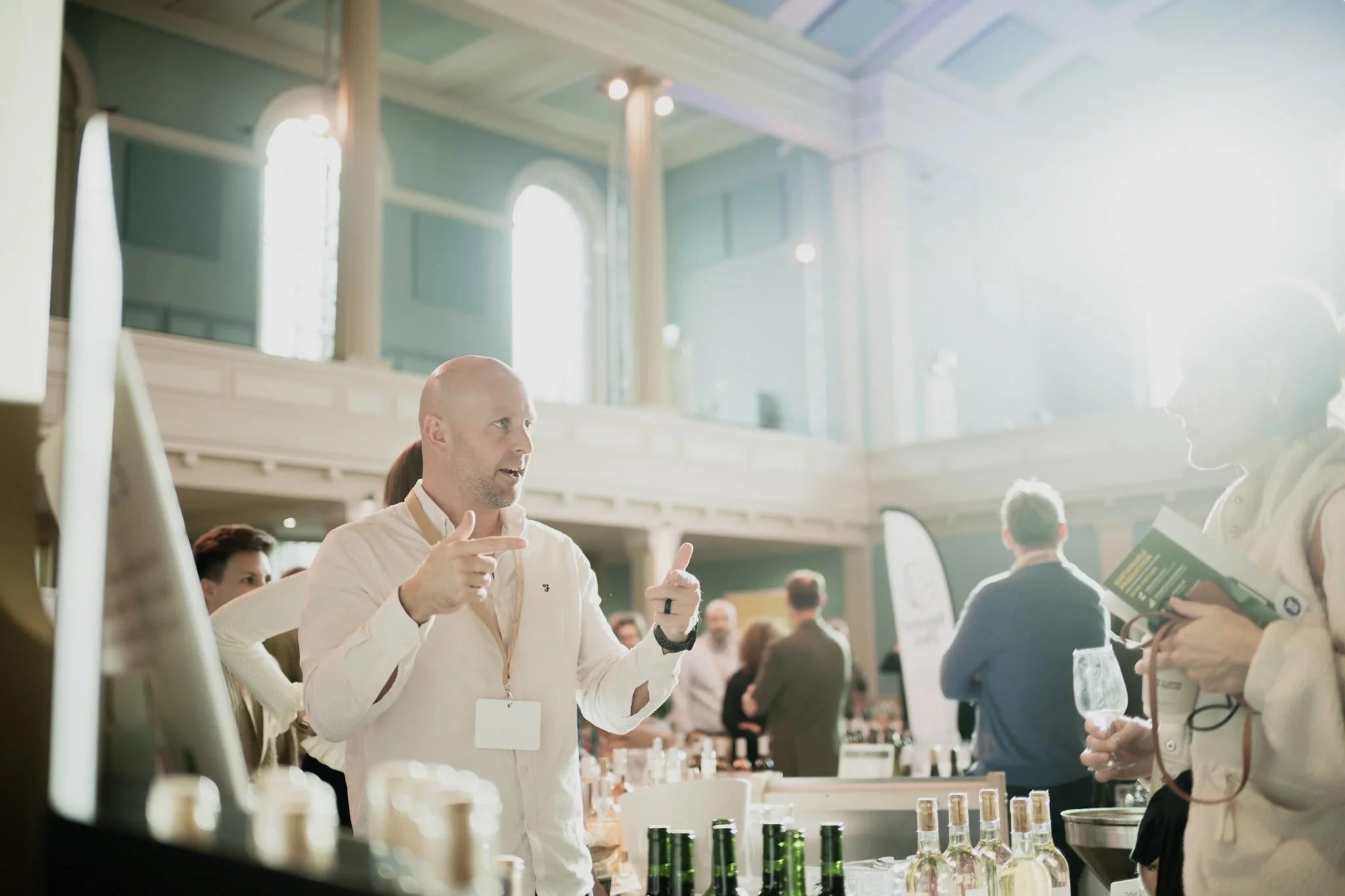 A man with a bald head and light-colored shirt talking and gesturing with his hands at a professional event or conference, with other attendees in the background, bottles on a table, and a bright, spacious room with high ceilings and large windows.