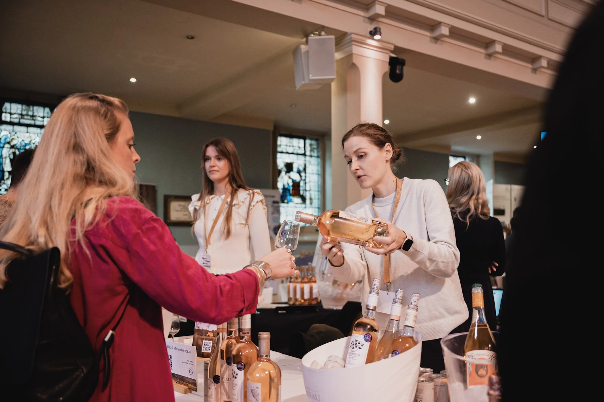 Women at a wine tasting event, with one woman pouring rosé wine into a glass and others observing, in a spacious room with stained glass windows.