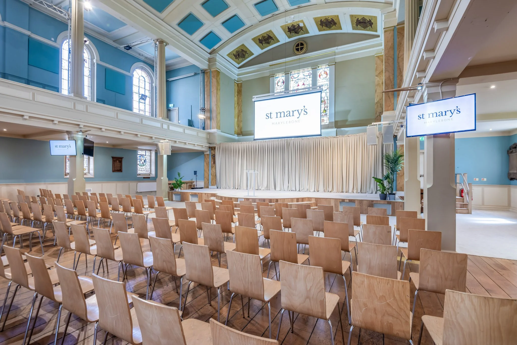 An empty church or auditorium with rows of wooden chairs facing a stage with a white curtain. There are multiple screens showing the logo 'st mary's MARYLEBONE' hanging above the stage. The room has a high, decorated ceiling with chandeliers, large stained glass windows, and pastel-colored walls.