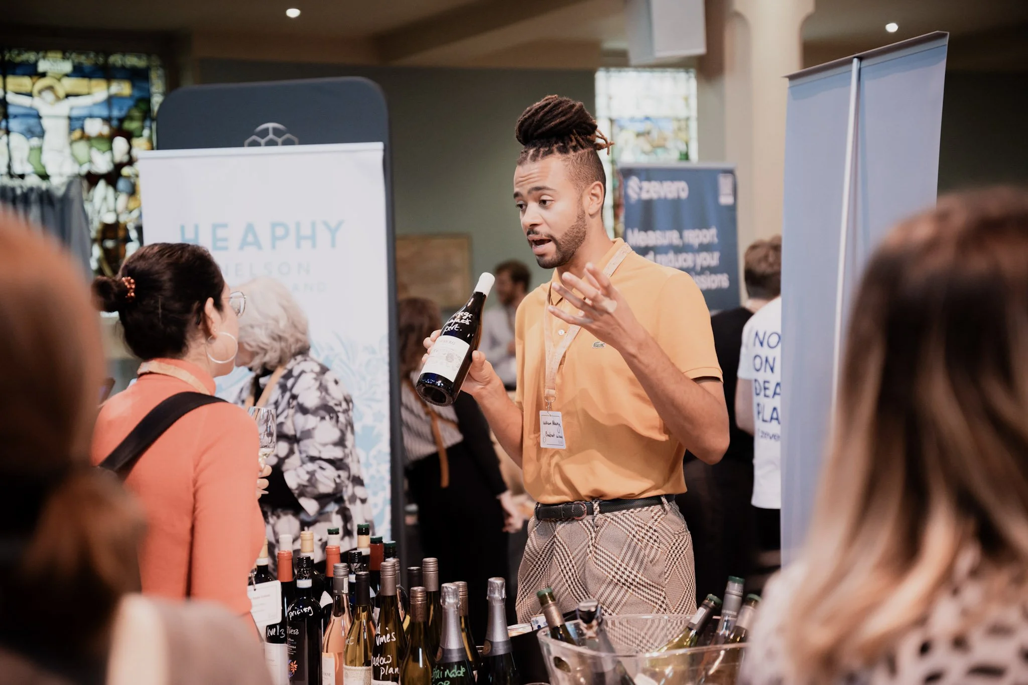 A man with dreadlocks presenting a wine bottle at a tasting event, surrounded by attendees and bottles of wine on display.