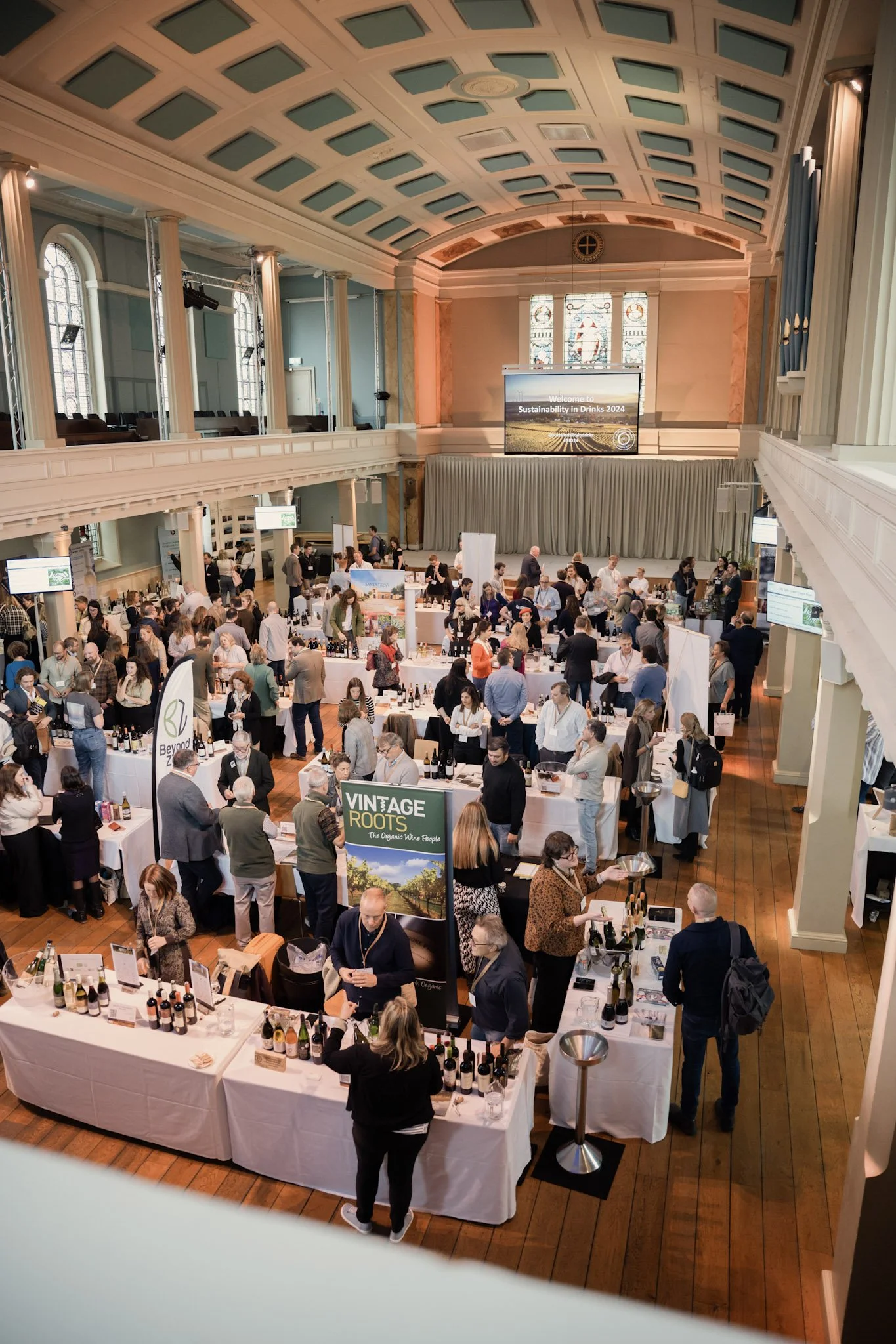 A large indoor event in a spacious hall with a high, decorated ceiling, filled with attendees browsing tables of wine bottles, with banners and screens displaying event information related to sustainability and vintage wines.