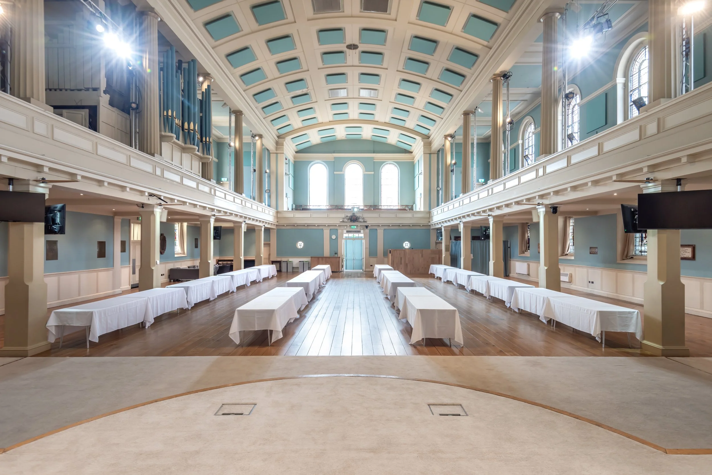 An elegant, spacious hall with high vaulted ceiling, large windows, and rows of tables covered with white cloth, set for an event or gathering.