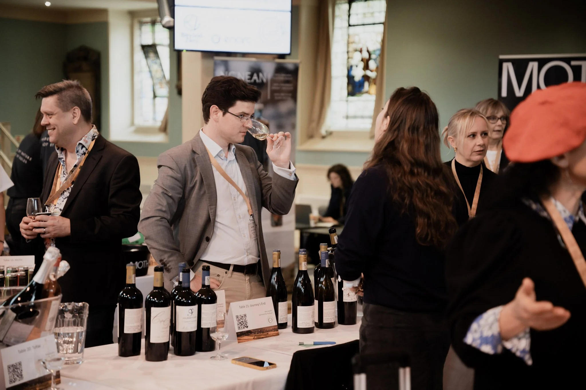 A man in a gray blazer tasting wine at a wine tasting event with several bottles of wine on display. Other people are engaged in conversations around him, and there are banners and a large screen in the background.