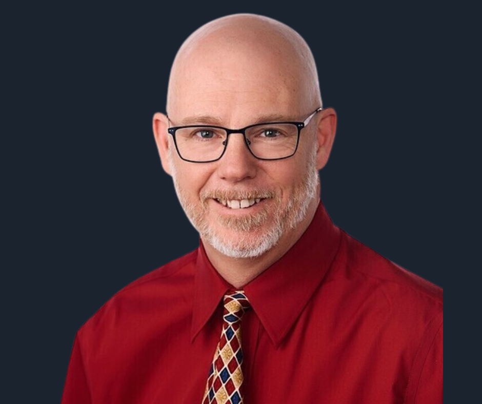 A smiling middle-aged man with glasses, a beard, and no hair, wearing a red shirt and a multicolored patterned tie against a white background.