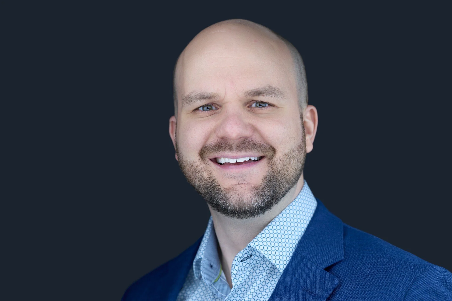 A smiling man with a beard wearing a light blue button-up shirt.