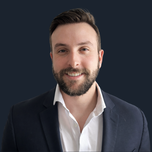 A portrait of a middle-aged man with a beard and bald head, wearing a black suit, white shirt, and a blue patterned tie.