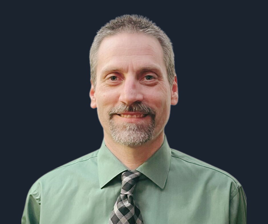 A middle-aged man with short, light brown hair and a beard, wearing a green dress shirt and a plaid tie, smiling against a plain white background.