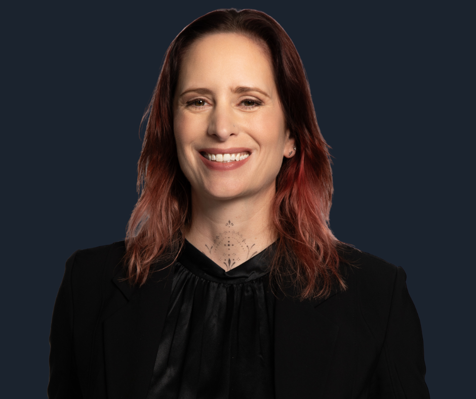 A woman with shoulder-length red hair, wearing a black blazer and matching top, smiling and holding her blazer lapels against a white background.