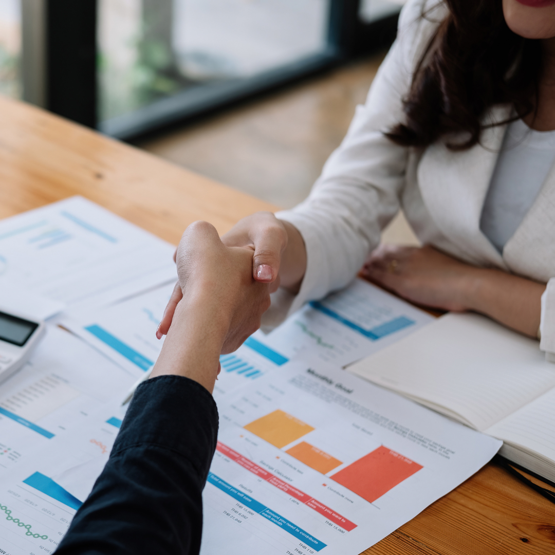 Two people shaking hands over a table with financial reports and documents during a business meeting.