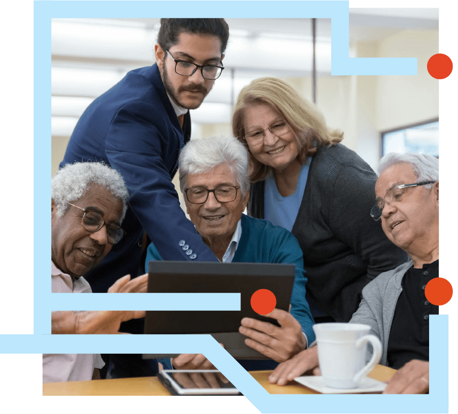 Group of five seniors and a young man gathered around a tablet, smiling and looking at the screen in a bright room with coffee cups on the table.