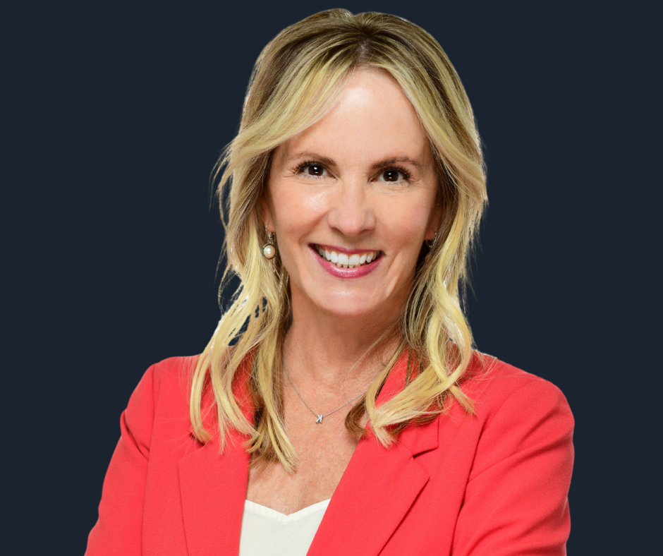 A woman with blonde hair, smiling, wearing a coral blazer, white blouse, and jewelry, posed with crossed arms against a white background.