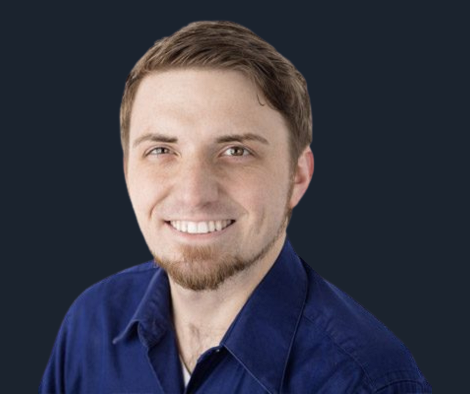 A young man with short brown hair and a beard, smiling, wearing a blue collared shirt against a white background.