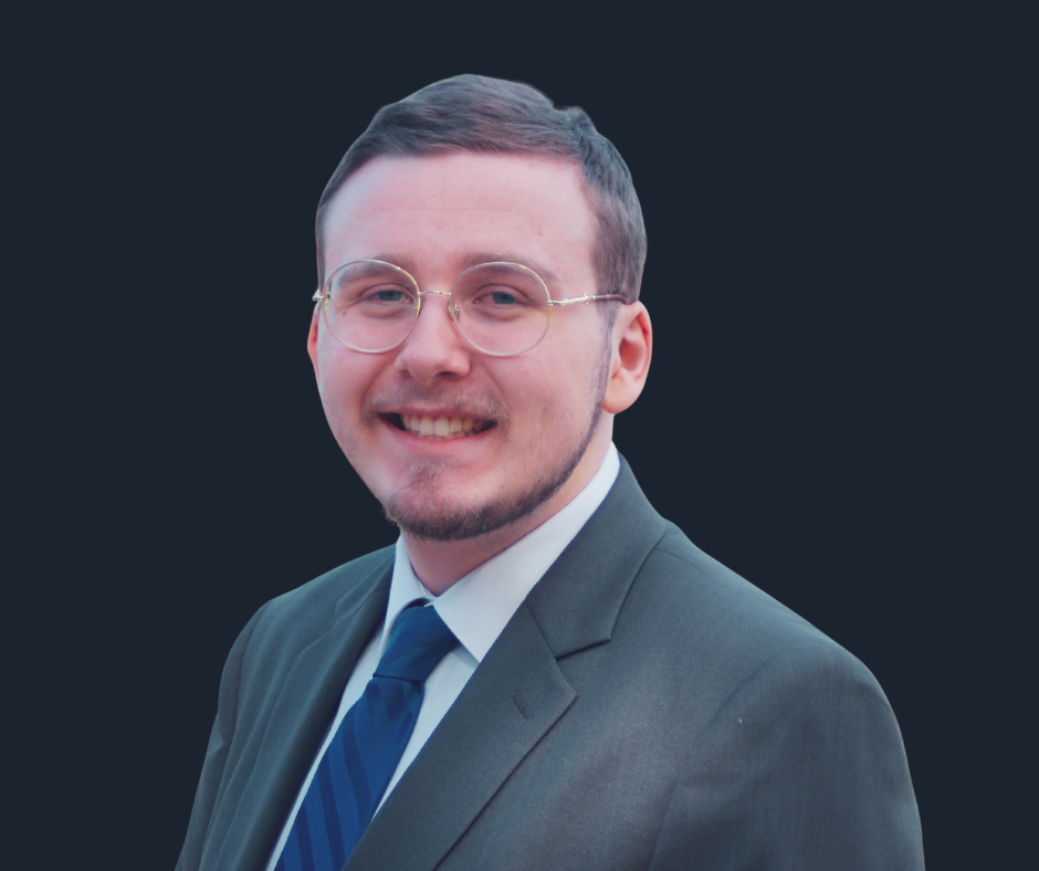 Headshot of a young man with short brown hair, glasses, wearing a dark suit, white shirt, and blue tie, smiling against a plain white background.