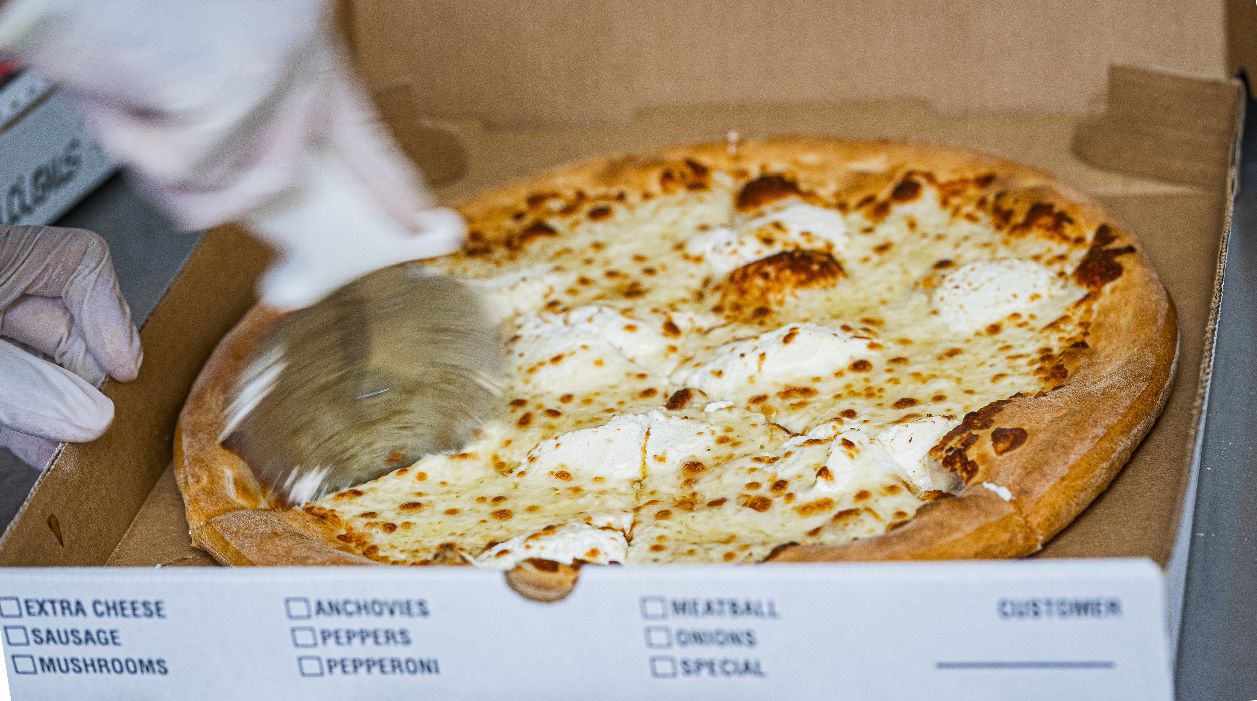 Person slicing a cheese pizza with a pizza cutter in a cardboard box.