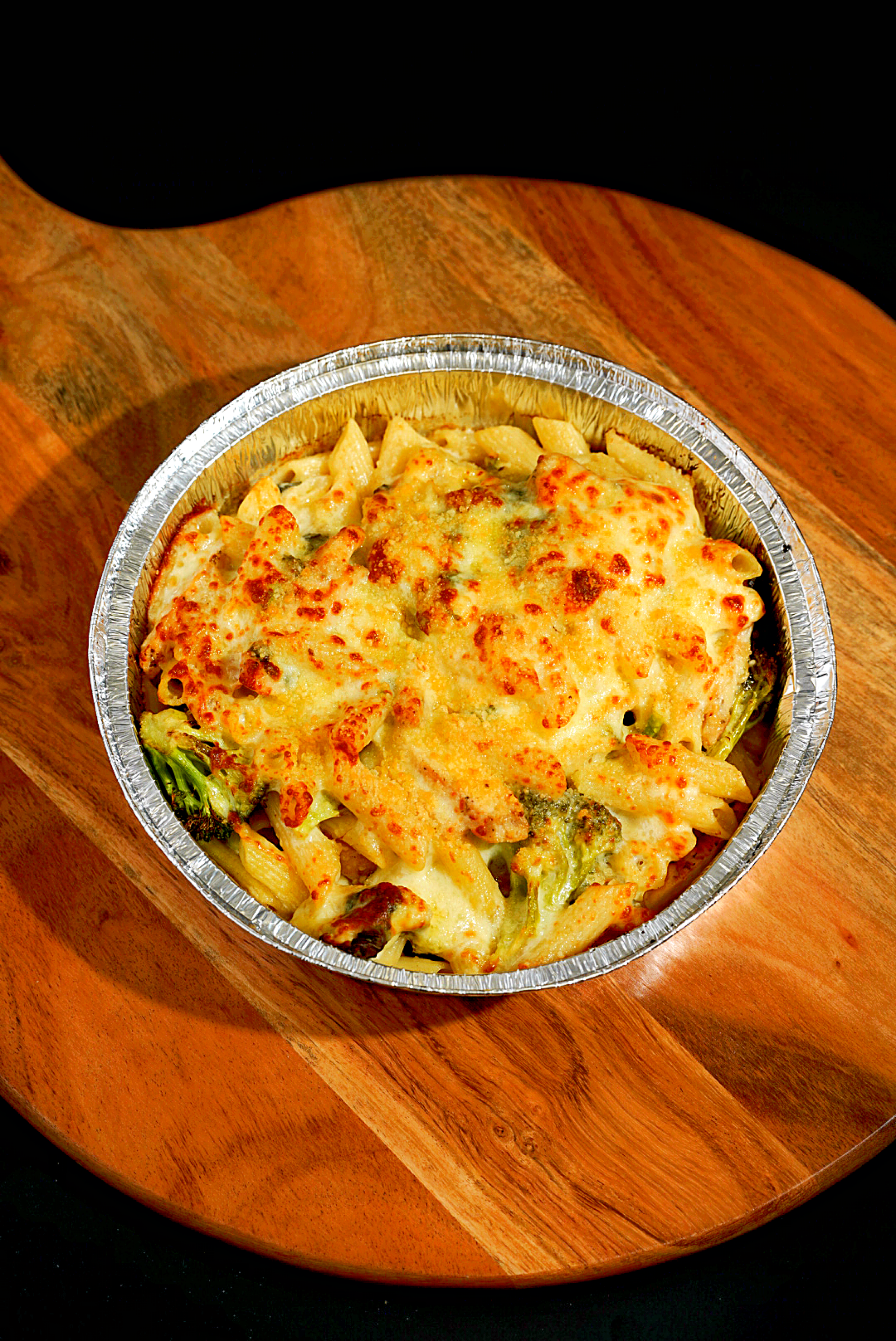 Baked cheesy pasta with broccoli in a foil container on a wooden board.