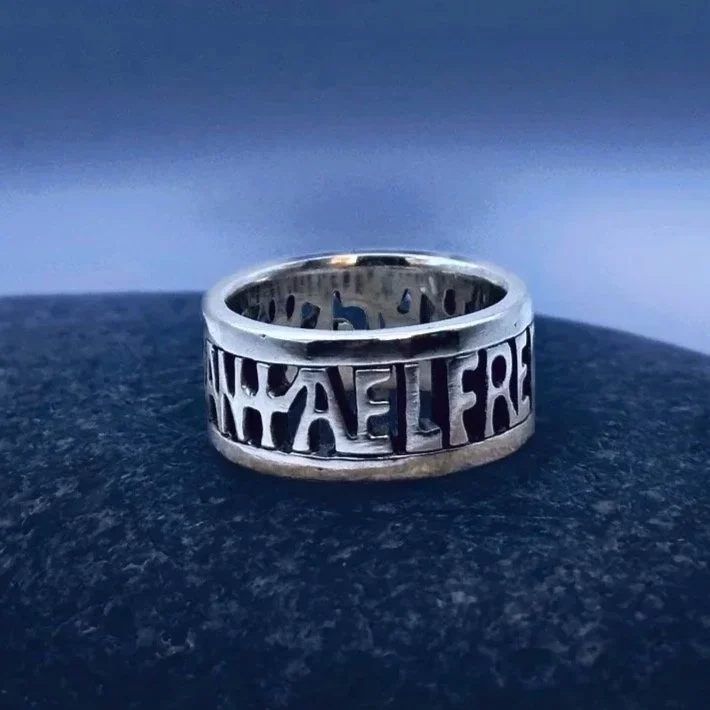 Close up of sterling silver Alfred the Great ring, inspired by The Alfred Jewel, which reads "Aelfred Mec Heht Gewyrcan" or Alfred Had Me Made, resting on top of a dark blue toned stone.