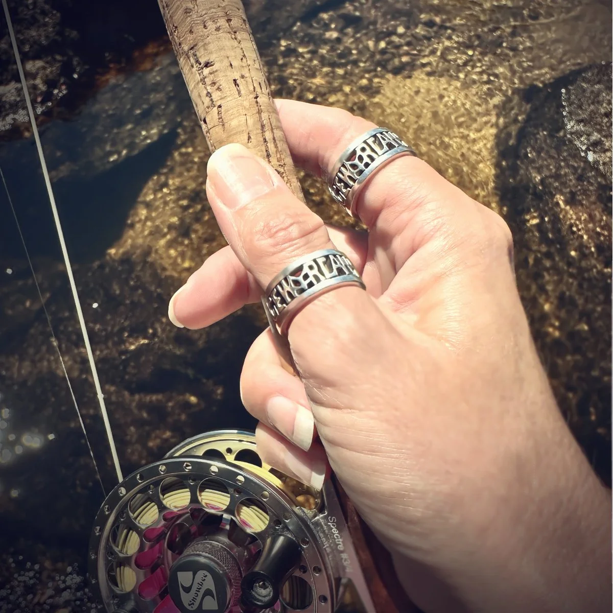 A person's hand holding a fishing rod with a reel, near a rocky water body. The person is wearing two silver rings with cut-out lettering.