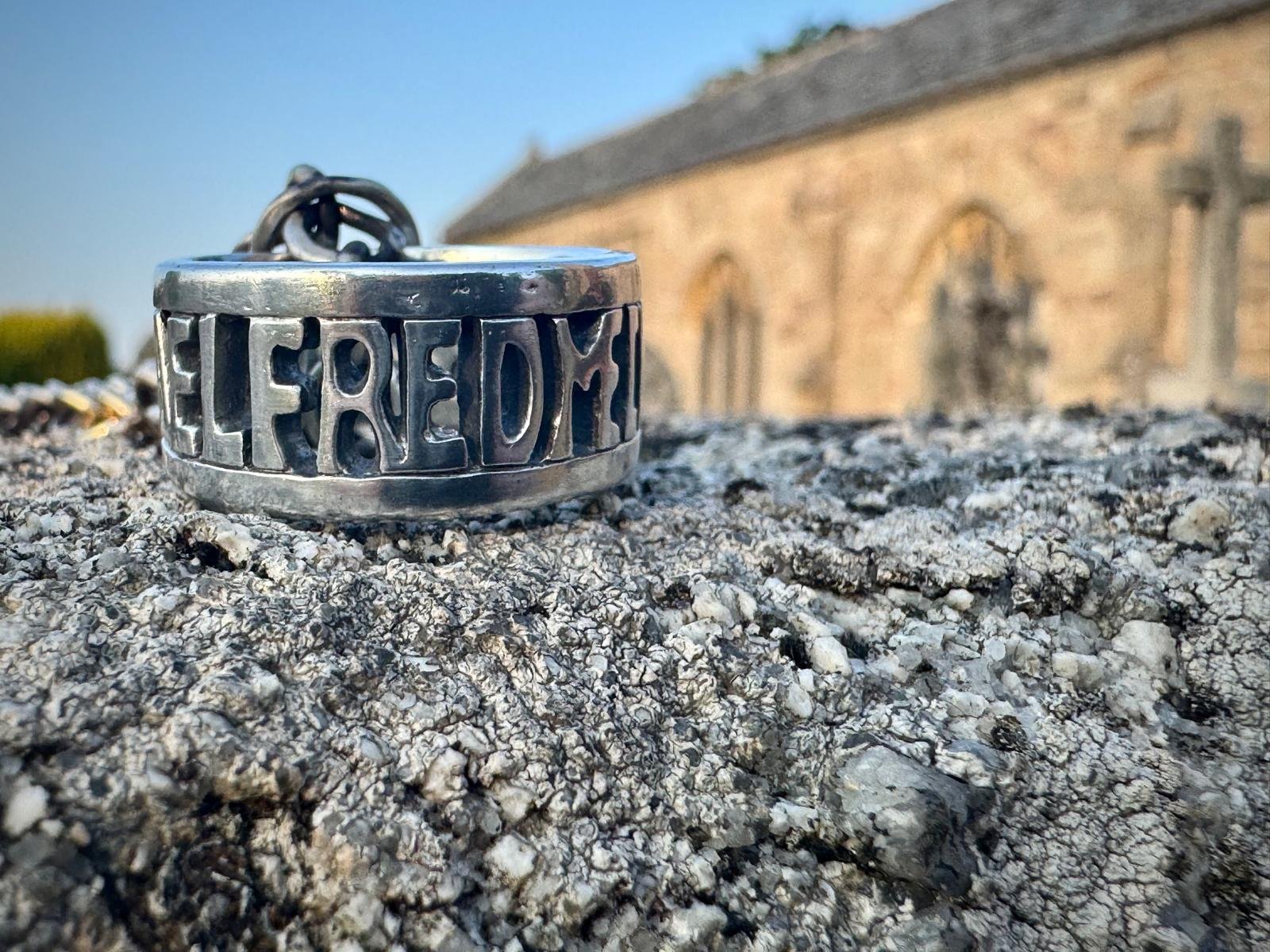 A close-up on a sterling silver Anglo Saxon and Vikings themed, Alfred the great ring reading "Aelfred Mec Heht Gewyrcan" or Alfred had me made, sitting onto of a tombstone with an ancient English church visible behind it and blue sky