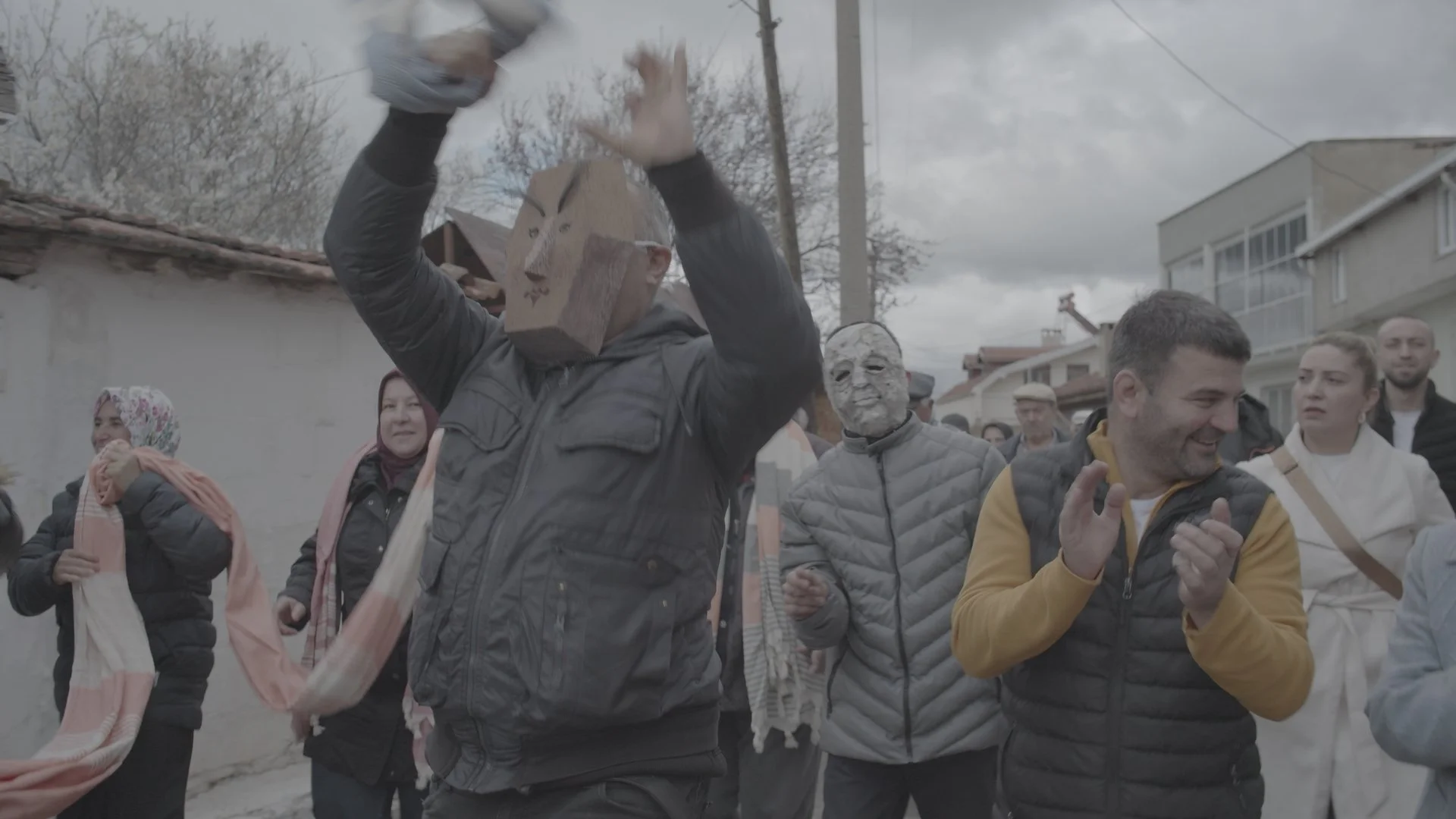 A group of people outdoors, some wearing masks, engaging in festivities or a celebration. One person wears a carved wooden mask and is dancing or performing, others are clapping or watching, under a cloudy sky.