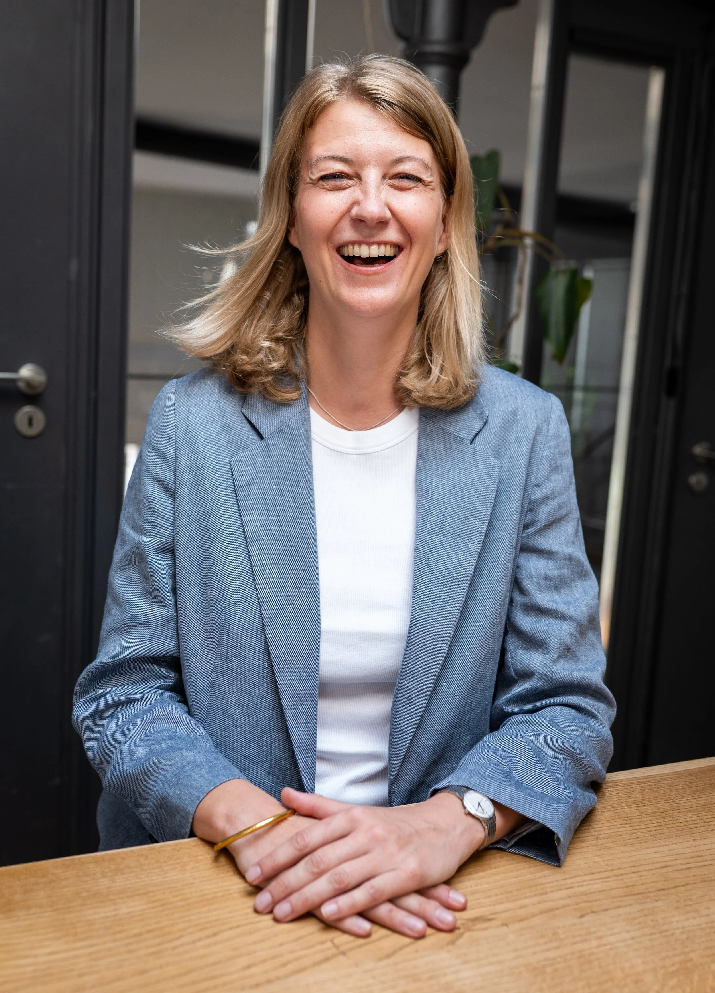 Frau lächelnd, sitzend an einem Holztisch, in einem Wohnzimmer oder Büro mit dunklen Wänden und Pflanzen im Hintergrund, trägt eine graue Blazer und weißes T-Shirt.