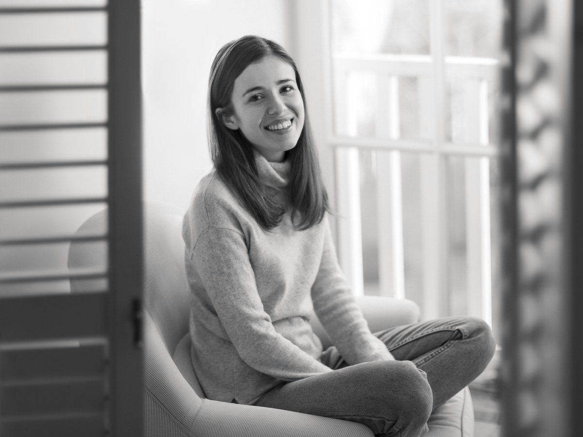 A young woman with shoulder-length hair smiling and sitting cross-legged on a sofa, seen through partially open window shutters.