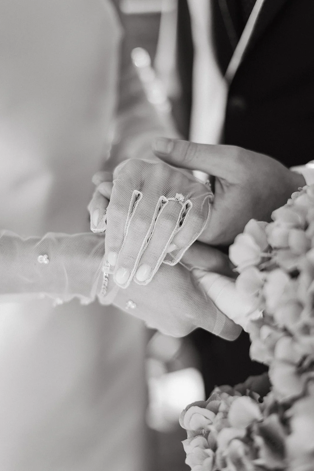 Close-up of a bride and groom's hands holding each other, with the bride's hand wearing a delicate lace glove and the groom's hand on top, holding her hand gently. The image is in black and white.