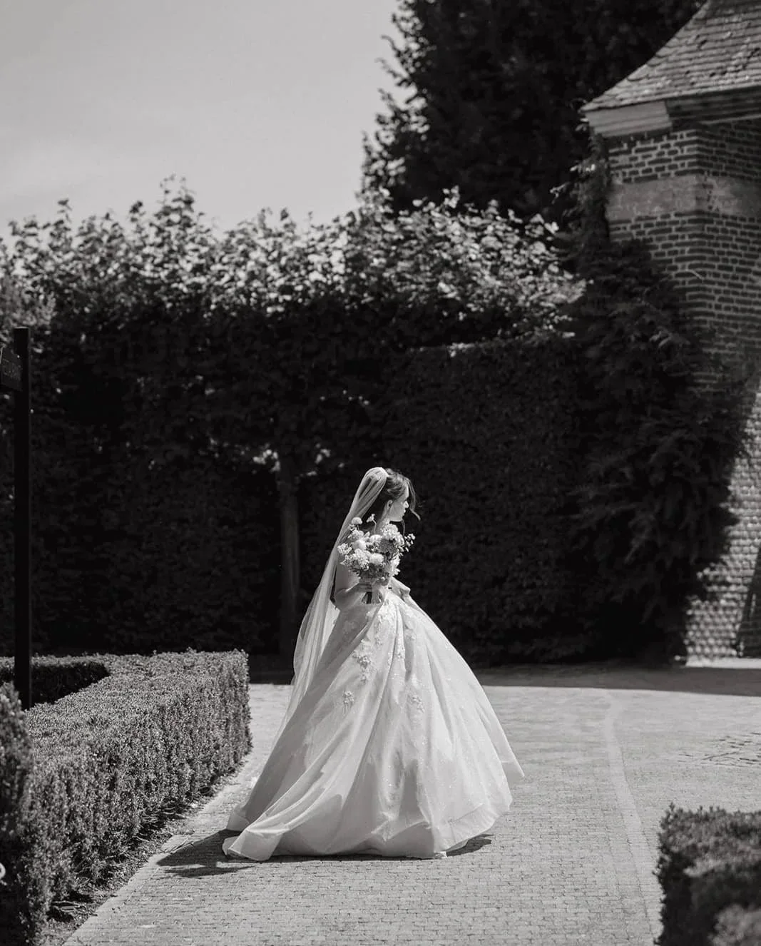 A bride in a wedding dress holding a bouquet of flowers walking on a paved path outdoors.