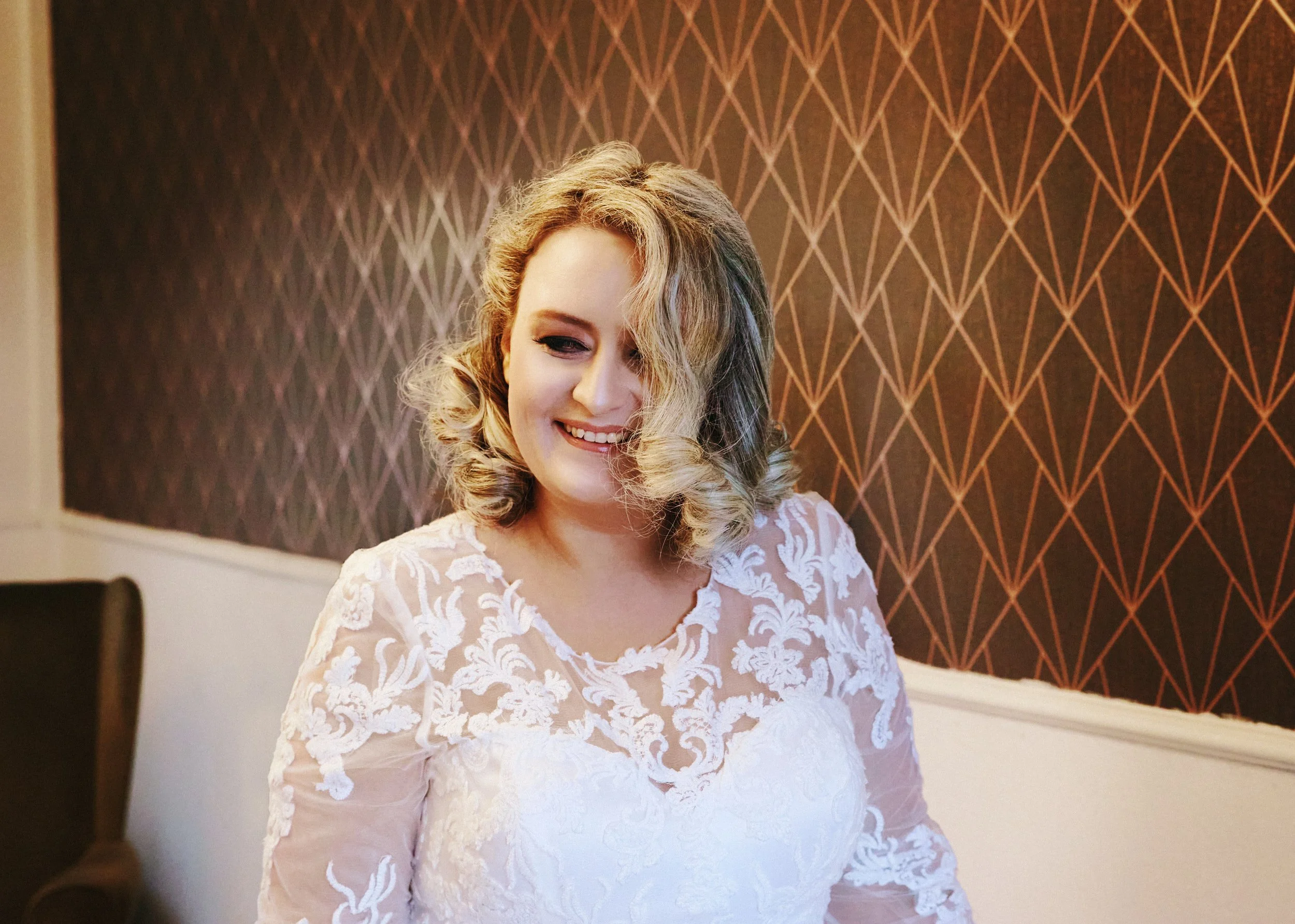 Woman in a white lace dress smiling with a geometric patterned wall background.
