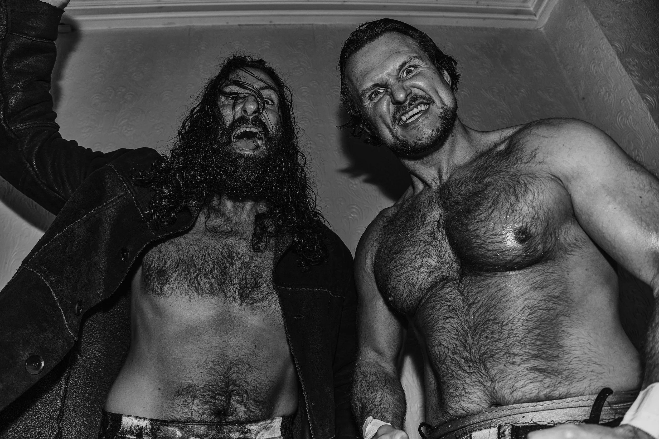 Two shirtless men with intense expressions, one with long hair and a beard, in a dramatic black and white setting.