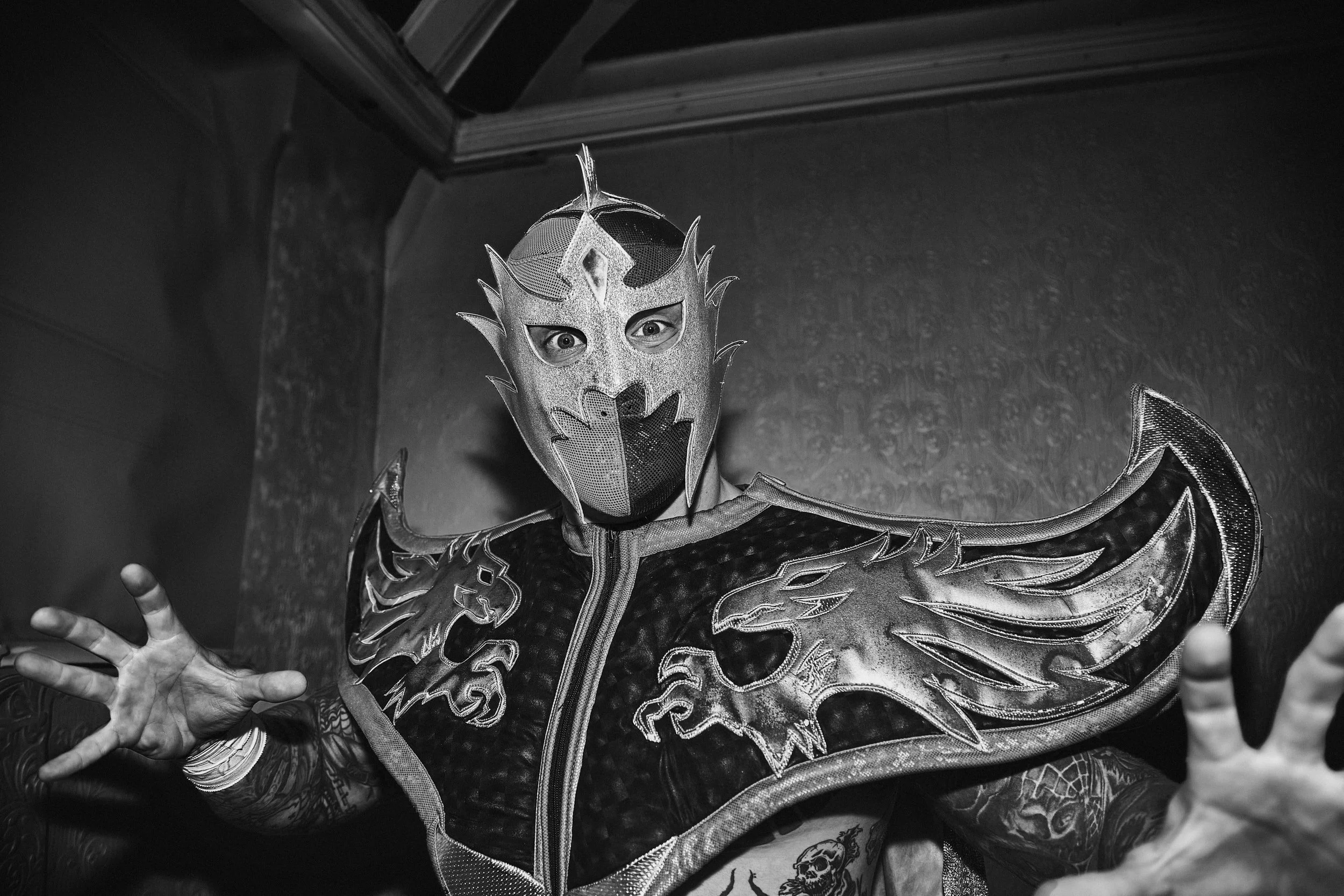 Black and white photo of a masked wrestler in an ornate costume with dragon designs, extending hands towards the camera.