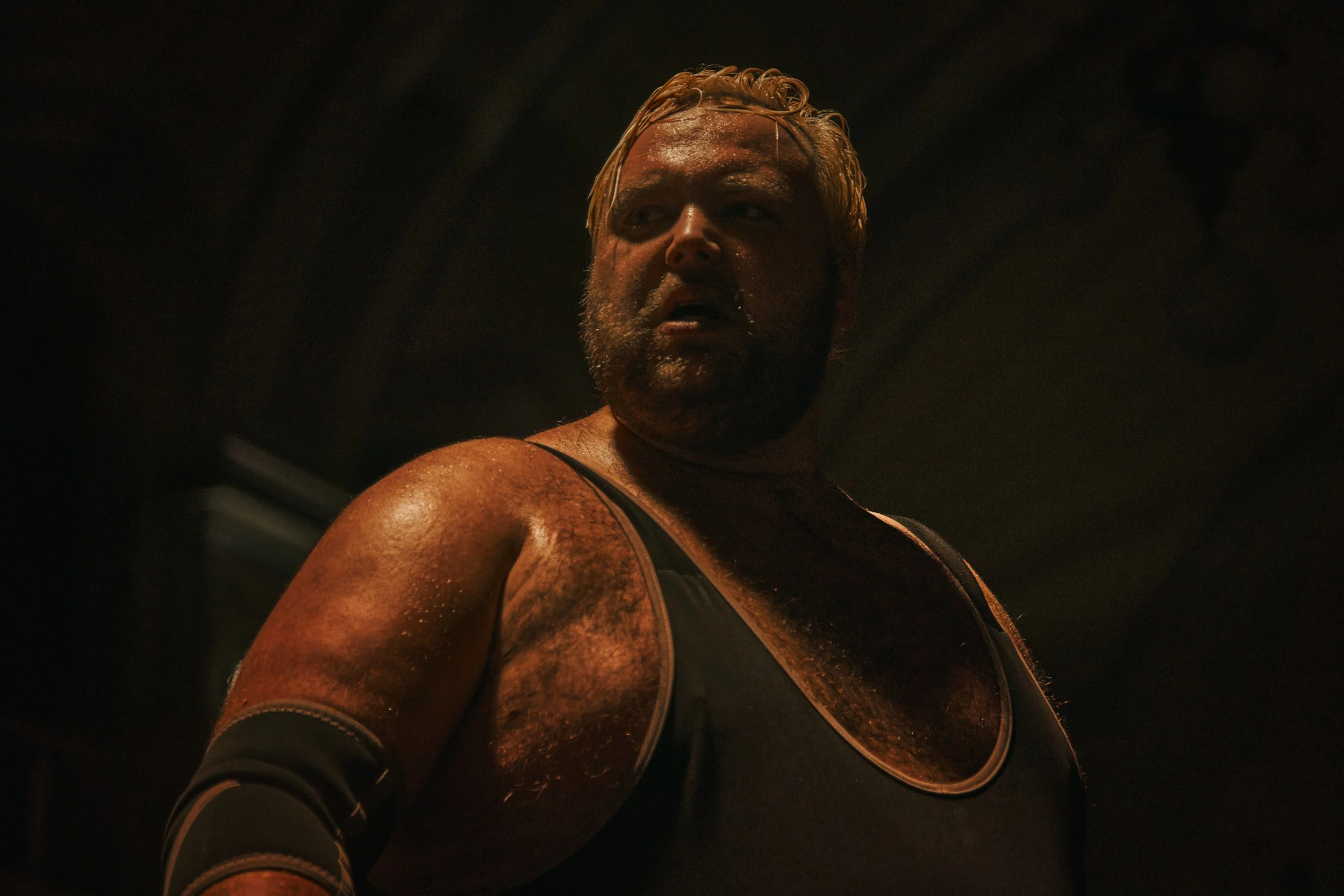 A wrestler with blonde hair and a beard, wearing a singlet and elbow pad, looking determinedly to the side in a dimly lit environment.