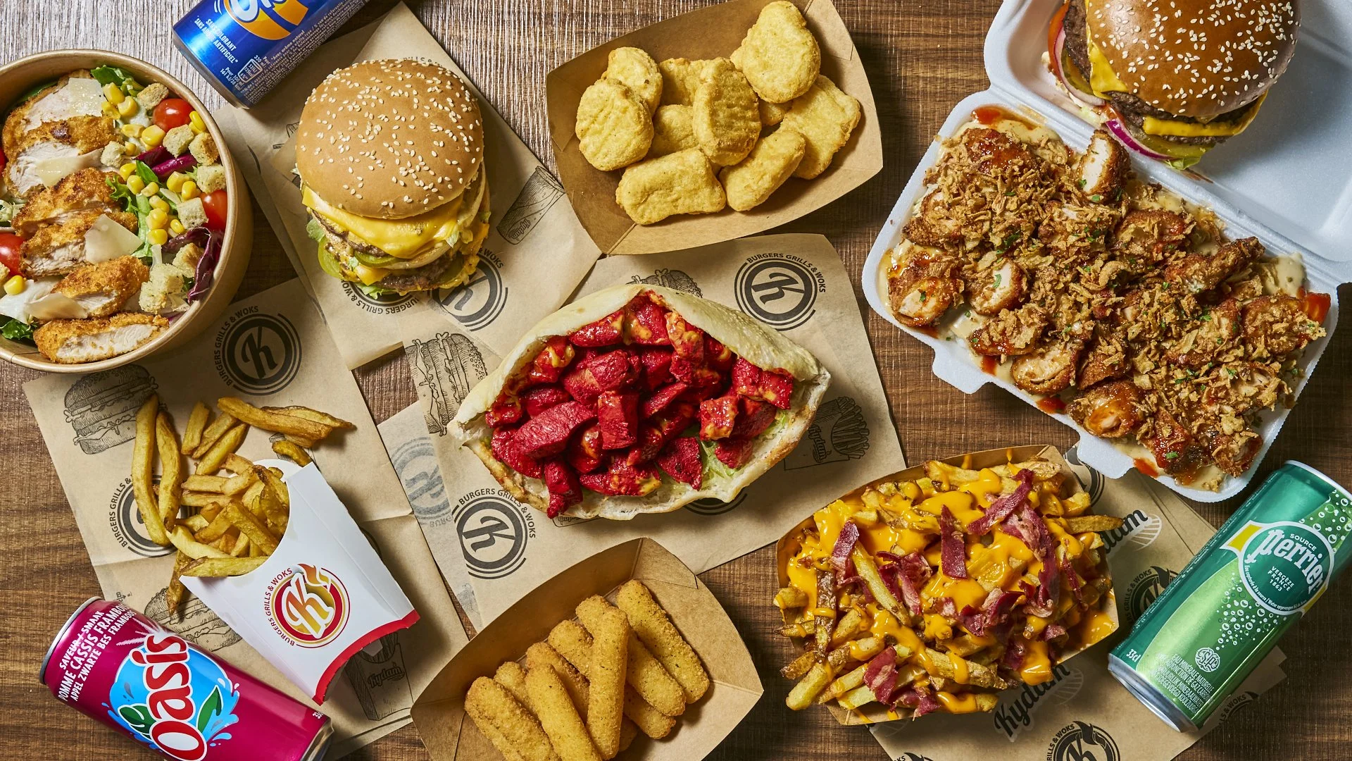 Repas fast-food comprenant plusieurs burgers, une salade, des frites, des soda, un sandwich chikka tandoori, des nuggets, et un crousty poulet avec des sauces sur une table en bois. Menus halal.