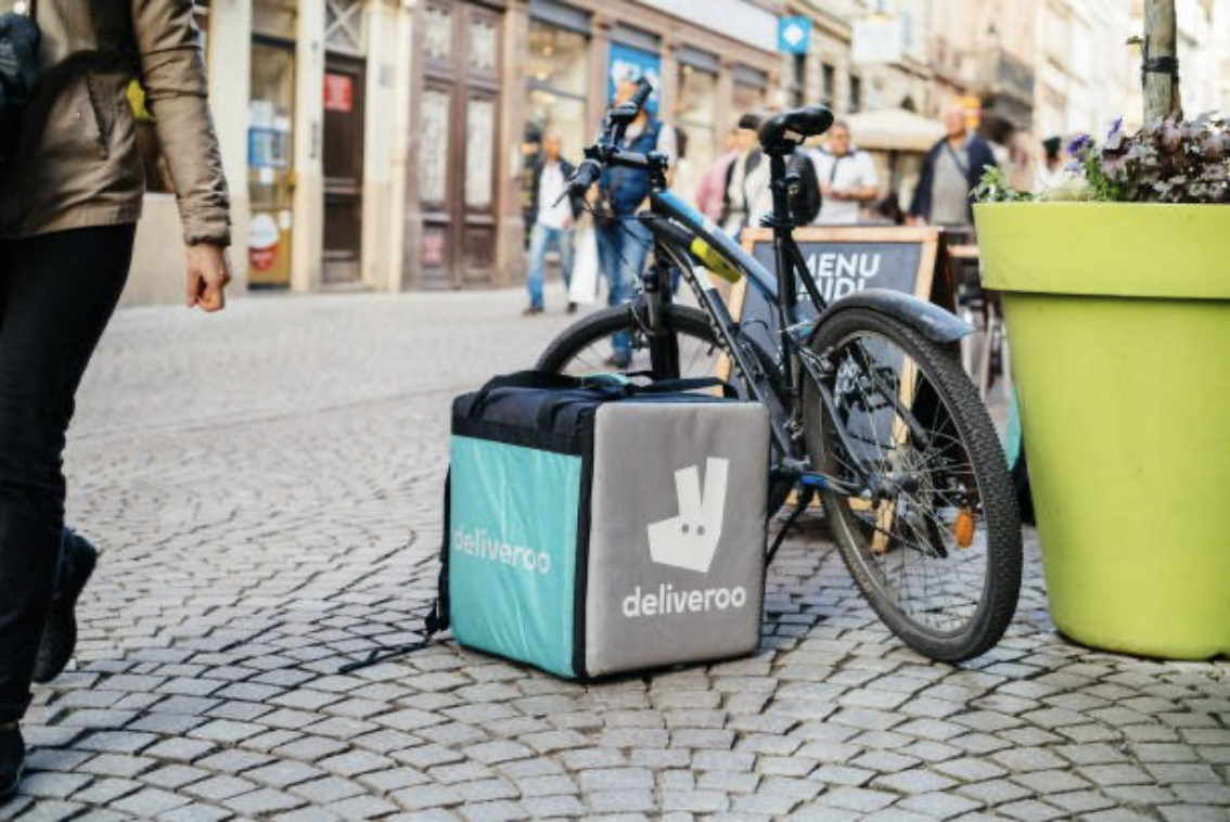 Un vélo de livraison avec une boîte de livraison 'deliveroo' attachée, stationné sur une rue pavée animée. Menus halal.