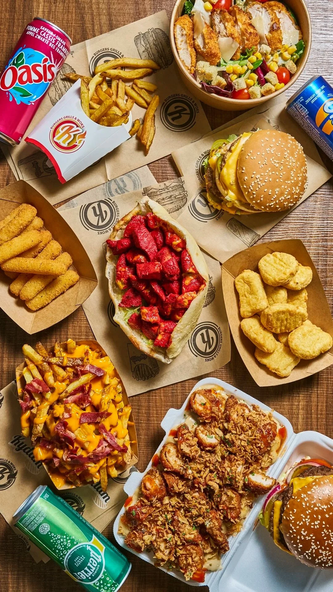 Repas fast-food comprenant plusieurs burgers, une salade, des frites, des soda, un sandwich chikka tandoori, des nuggets, et un crousty poulet avec des sauces sur une table en bois. Menus halal.