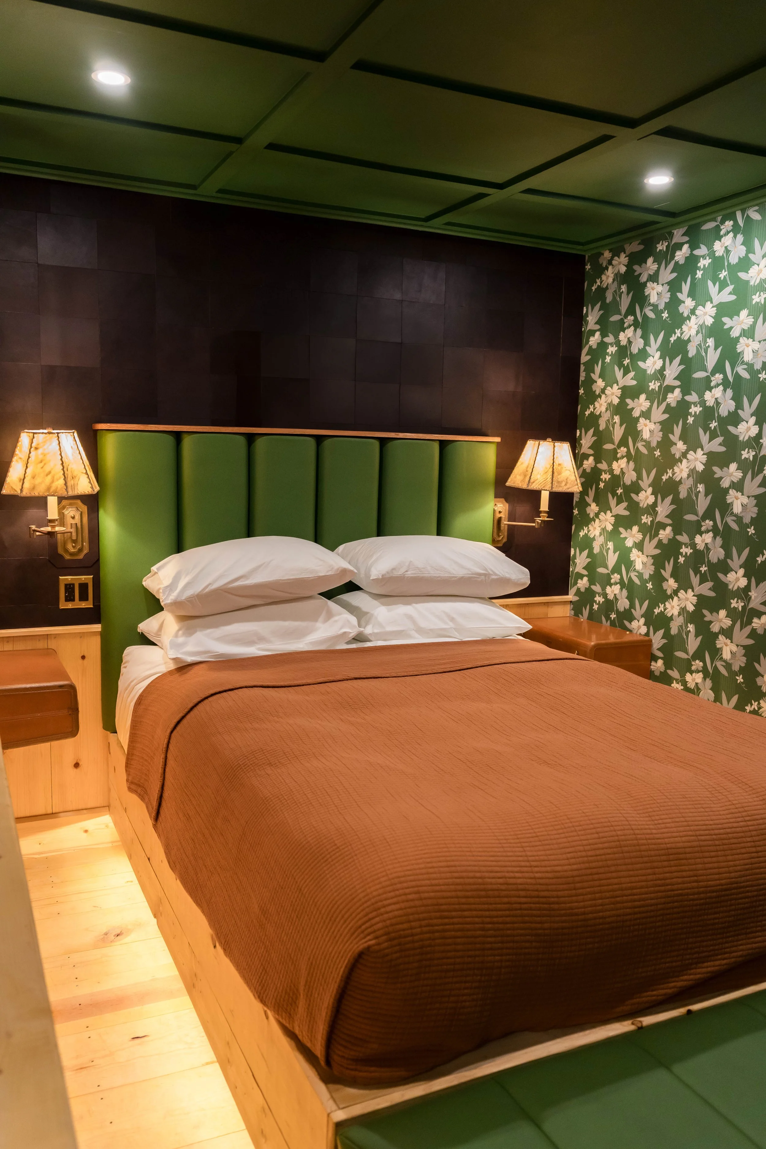 A hotel room with a bed that has a green upholstered headboard, six pillows, a brown bedspread, and two bedside lamps on a wooden nightstand, with dark wall and floral-patterned wallpaper