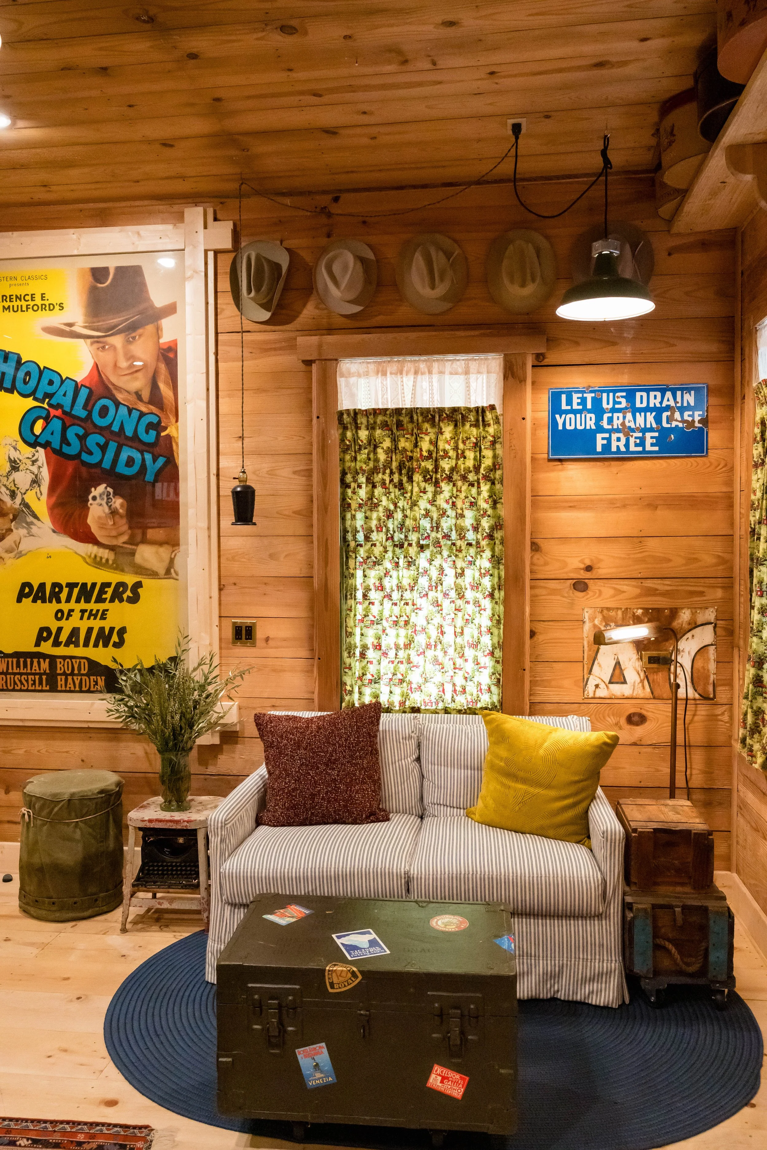 Cozy rustic living room with wood-paneled walls, a striped sofa with red and yellow pillows, a blue oval rug, vintage suitcases, a vintage style lamp, and a window with floral curtains. There are movie posters, hats on the wall, and a sign that reads "Let us drain your crank case free."