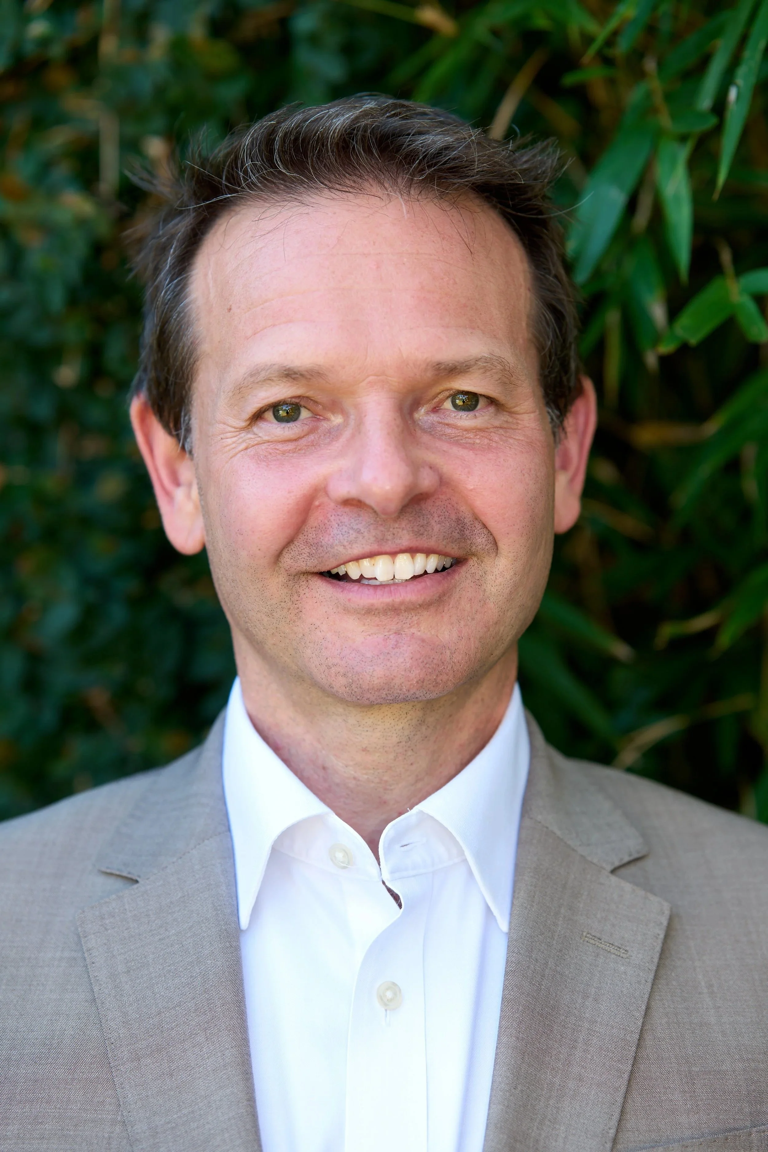 Dr Christoph Langenberg, smiling in a white dress shirt and beige blazer standing in front of green foliage.
