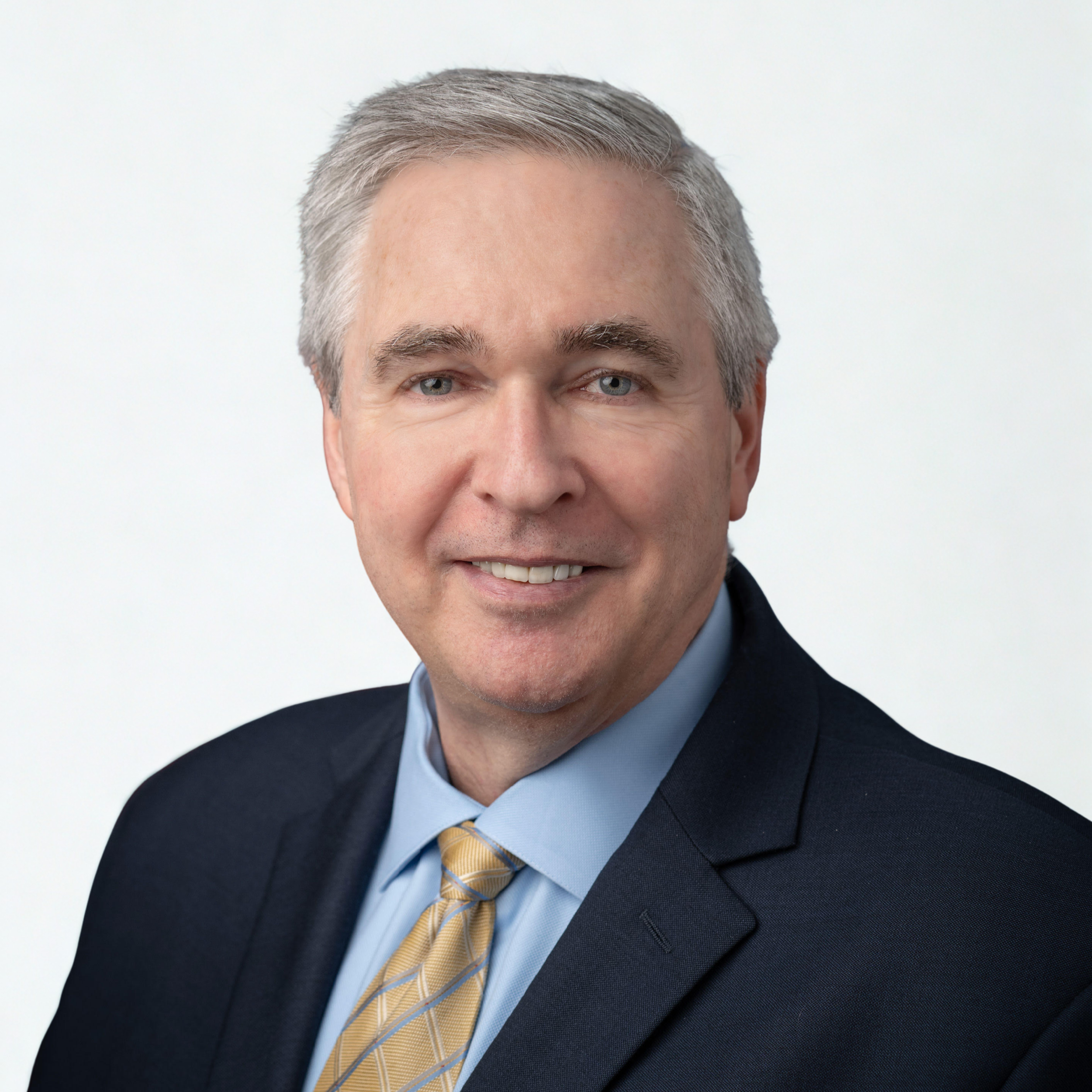 A professional headshot of a middle-aged man with gray hair, wearing a dark suit, light blue shirt, and yellow tie, smiling against a plain white background.
