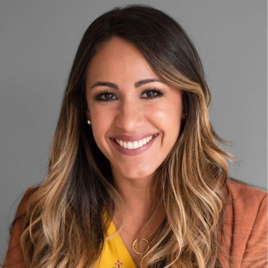 Smiling woman with long wavy hair, wearing a yellow top, brown jacket, and a delicate necklace, against a gray background.