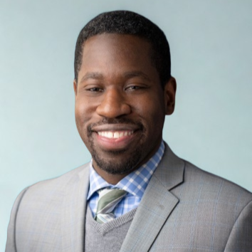 Portrait of a smiling African American man in a gray suit with a plaid shirt and tie, against a light blue background.