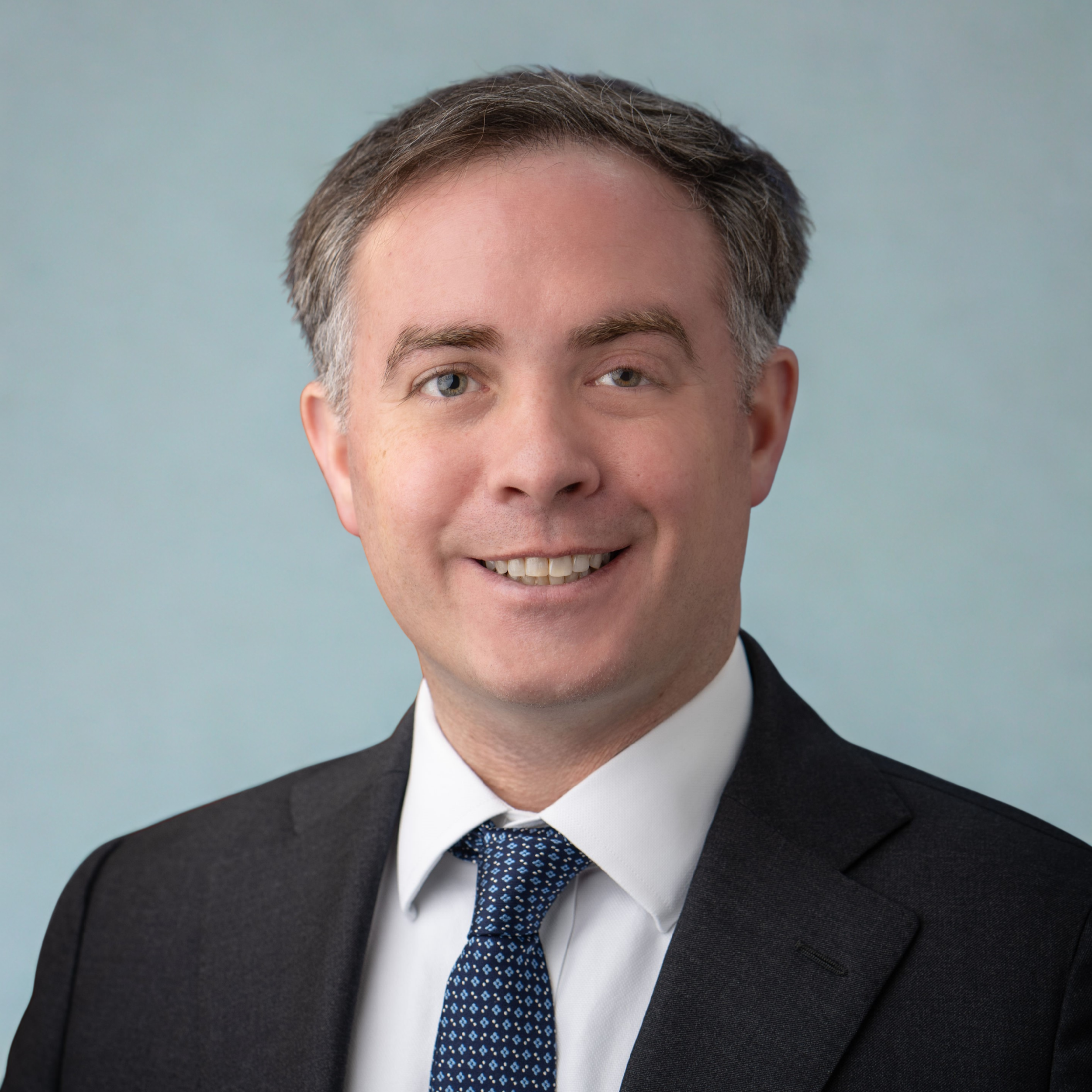 Professional headshot of a smiling man in a suit and tie against a light background.