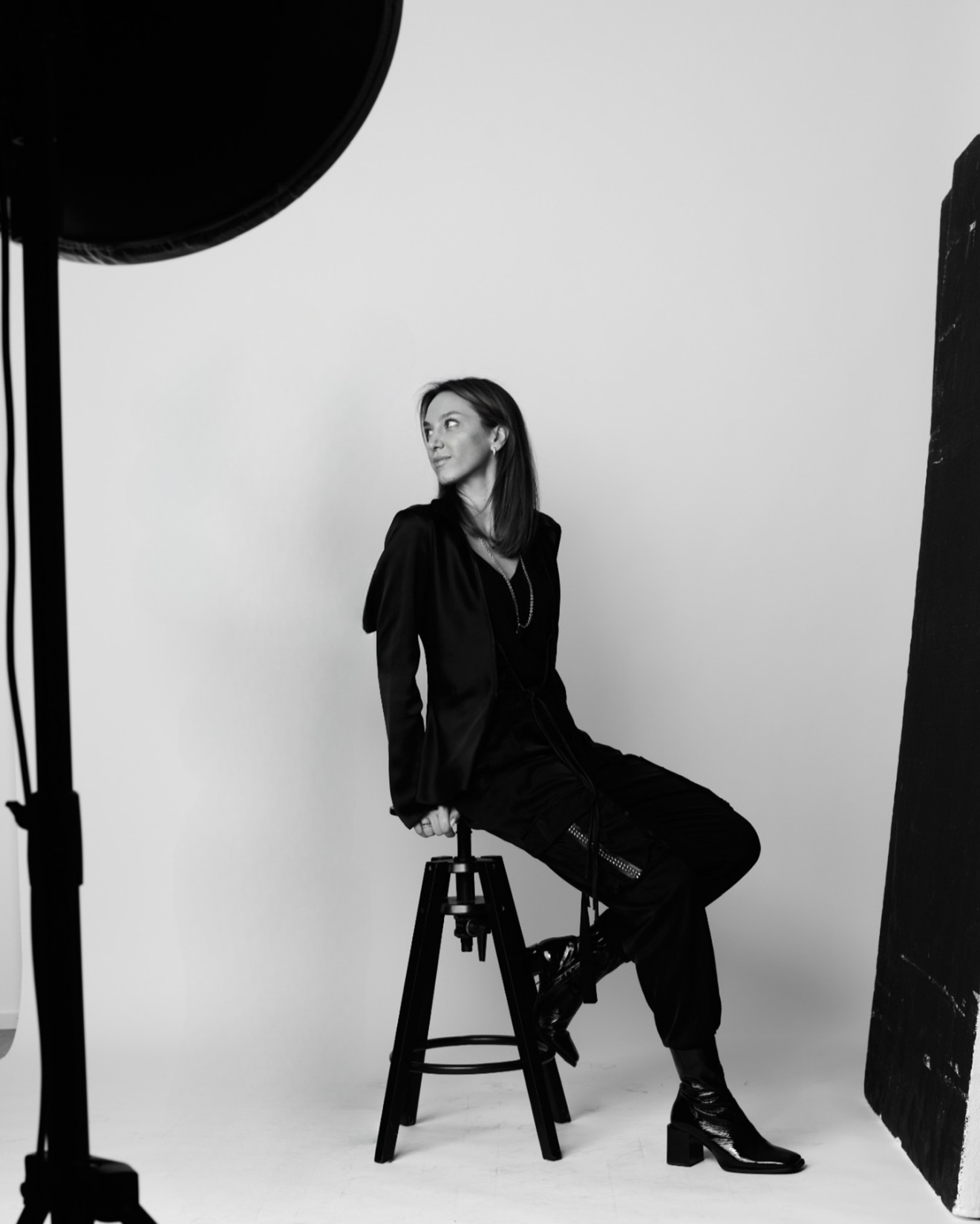 Black and white photo of a woman sitting on a stool in a photo studio with a plain background, looking to the side.