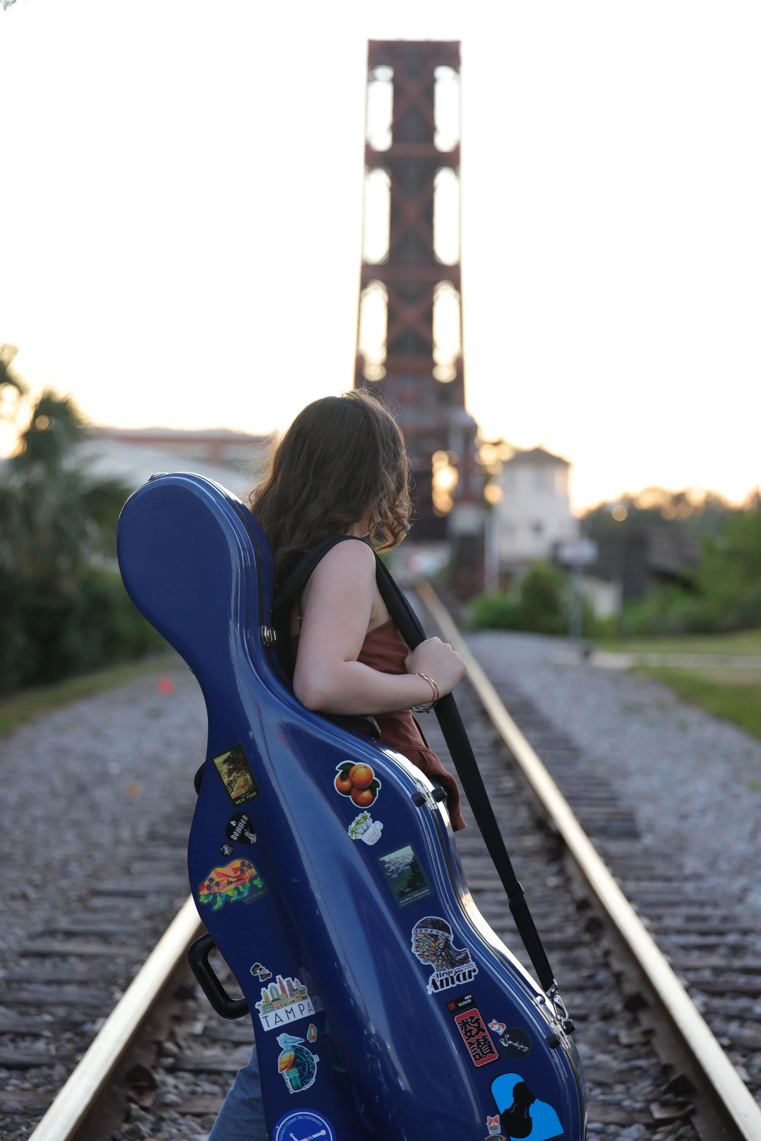 Susan Lindemann carrying a blue cello case with stickers, walking along train tracks at sunset, with a tall metal structure in the background.