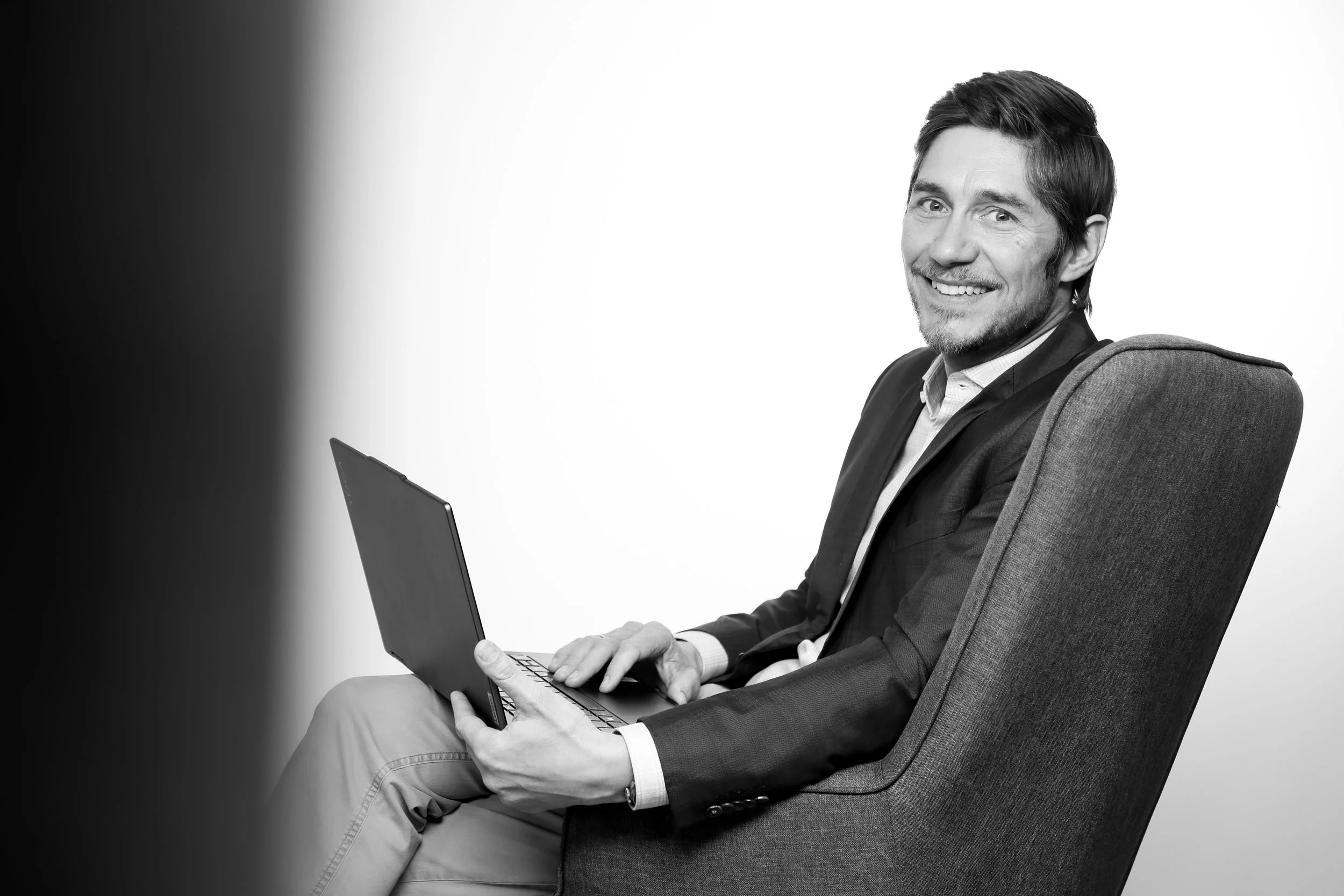 Black and white photo of a smiling man sitting in an armchair with a laptop in his hands.