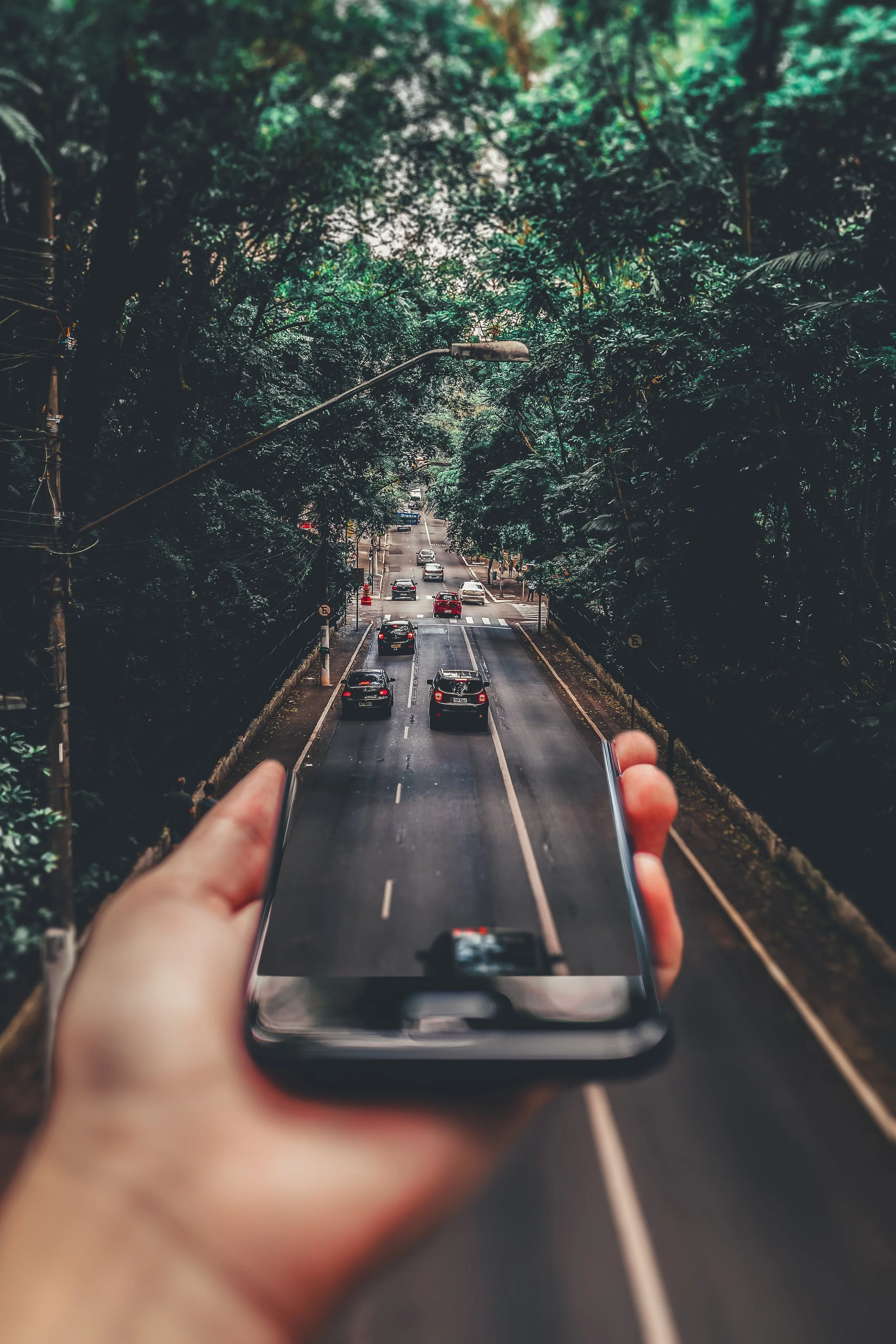 A person's hand holding a smartphone, with the camera view on the phone showing a busy street with cars and trees, creating an illusion of a mini street inside the phone.