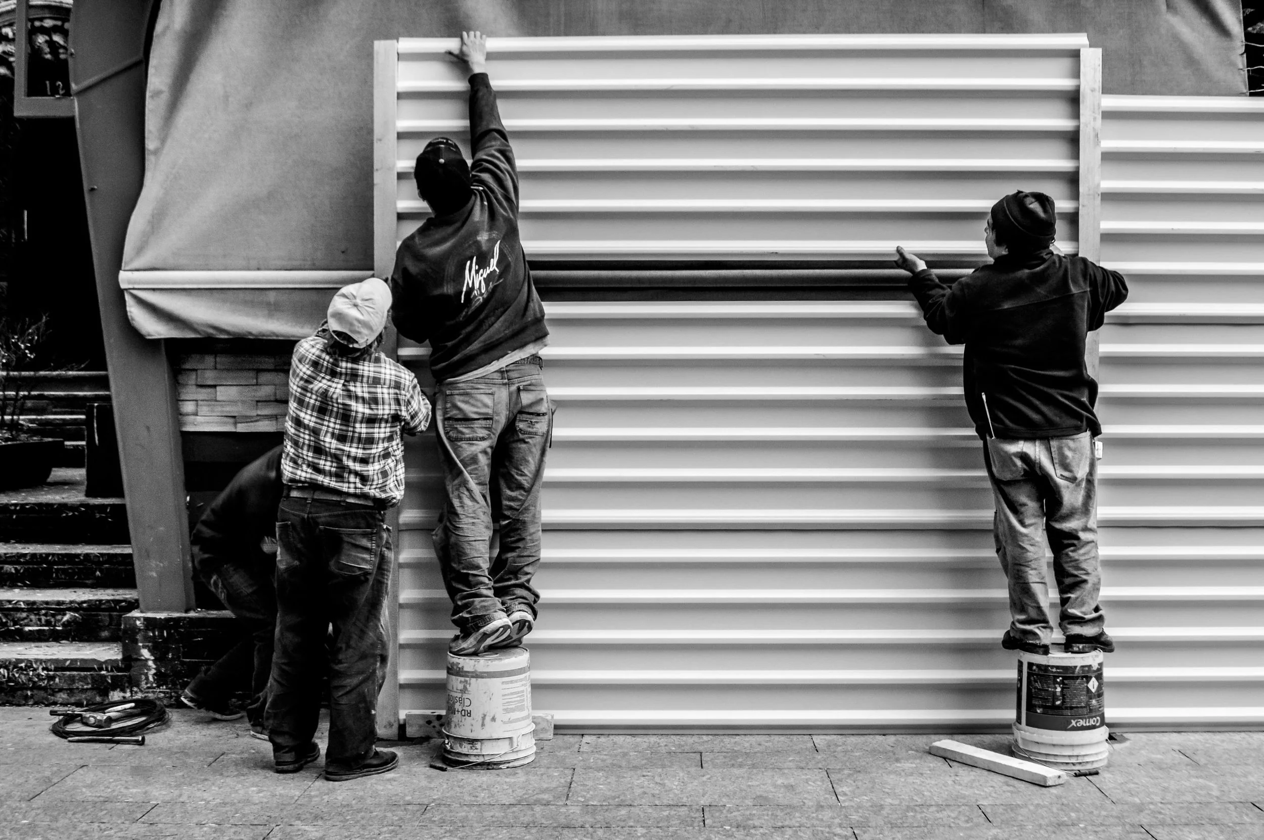 Four people working together on shutter installation, with two standing on paint cans, on a building exterior.