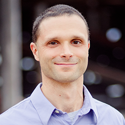 A man with short dark hair and light skin smiling, wearing a light blue collared shirt, outdoors in daytime.